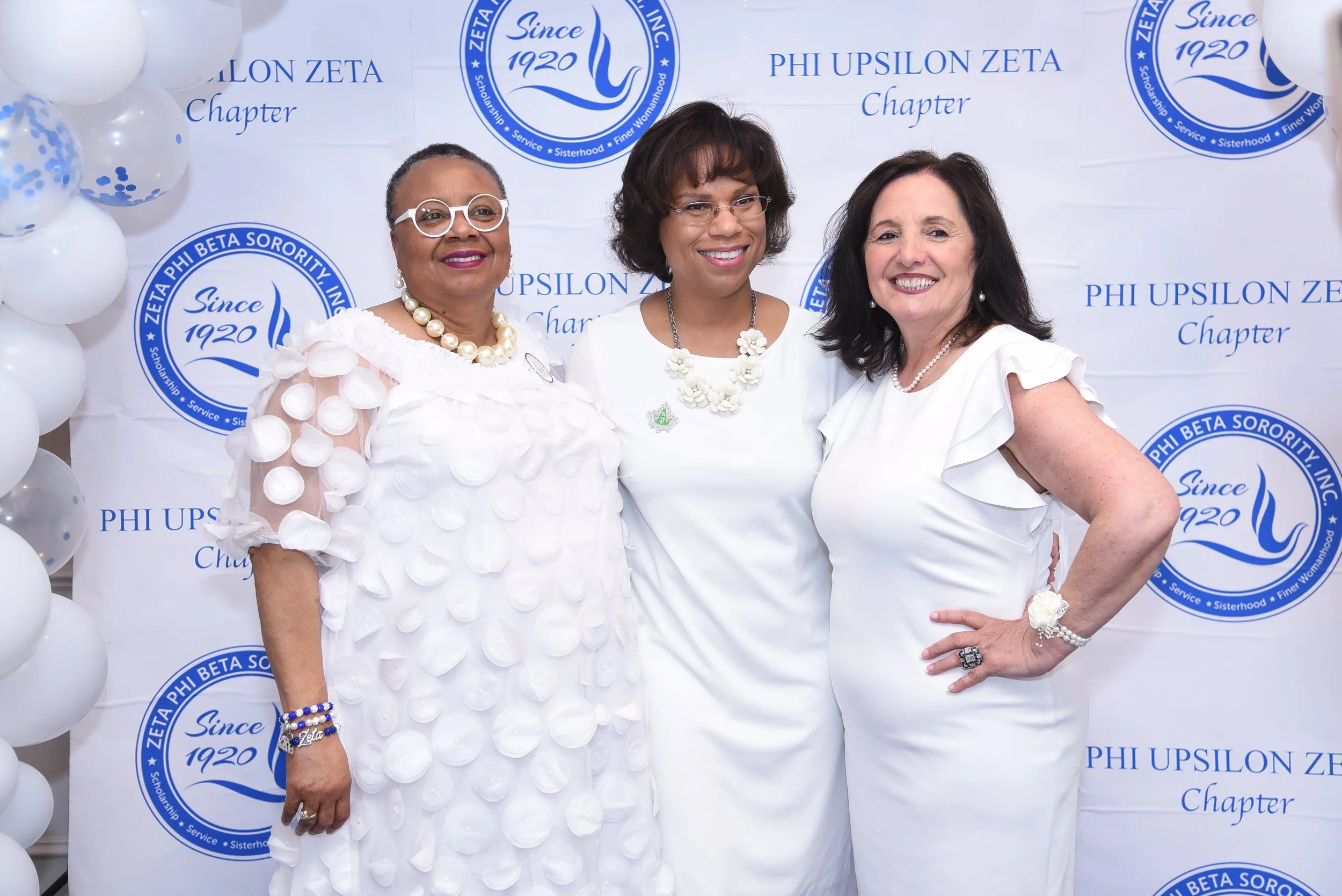 Three women standing together at a Phi Upsilon Zeta chapter event. They are wearing white outfits and smiling. The background features a banner with the Phi Upsilon Zeta logo, which includes the text "Since 1920". There are also white balloons with b
