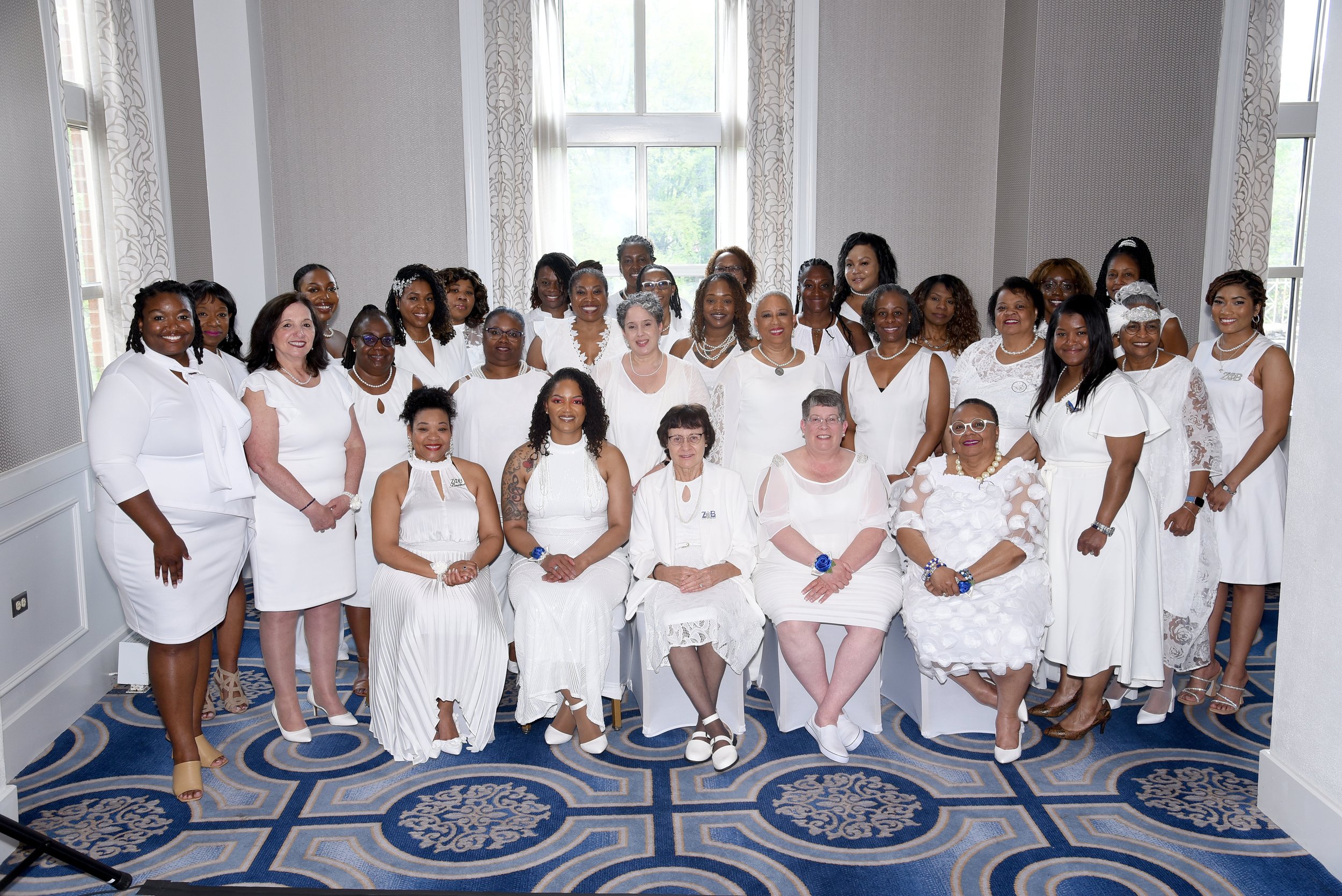 A group of women dressed in white posing for a photo in a room with large windows and patterned carpet.
