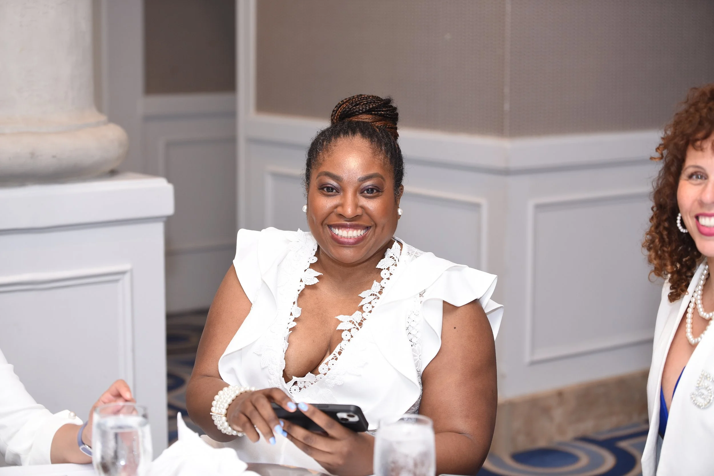 A woman with a braided bun hairstyle smiling, wearing a white dress with lace details, sitting at a table with glasses of water and using a smartphone. Another woman with curly hair and a pearl necklace is partially visible on the right.