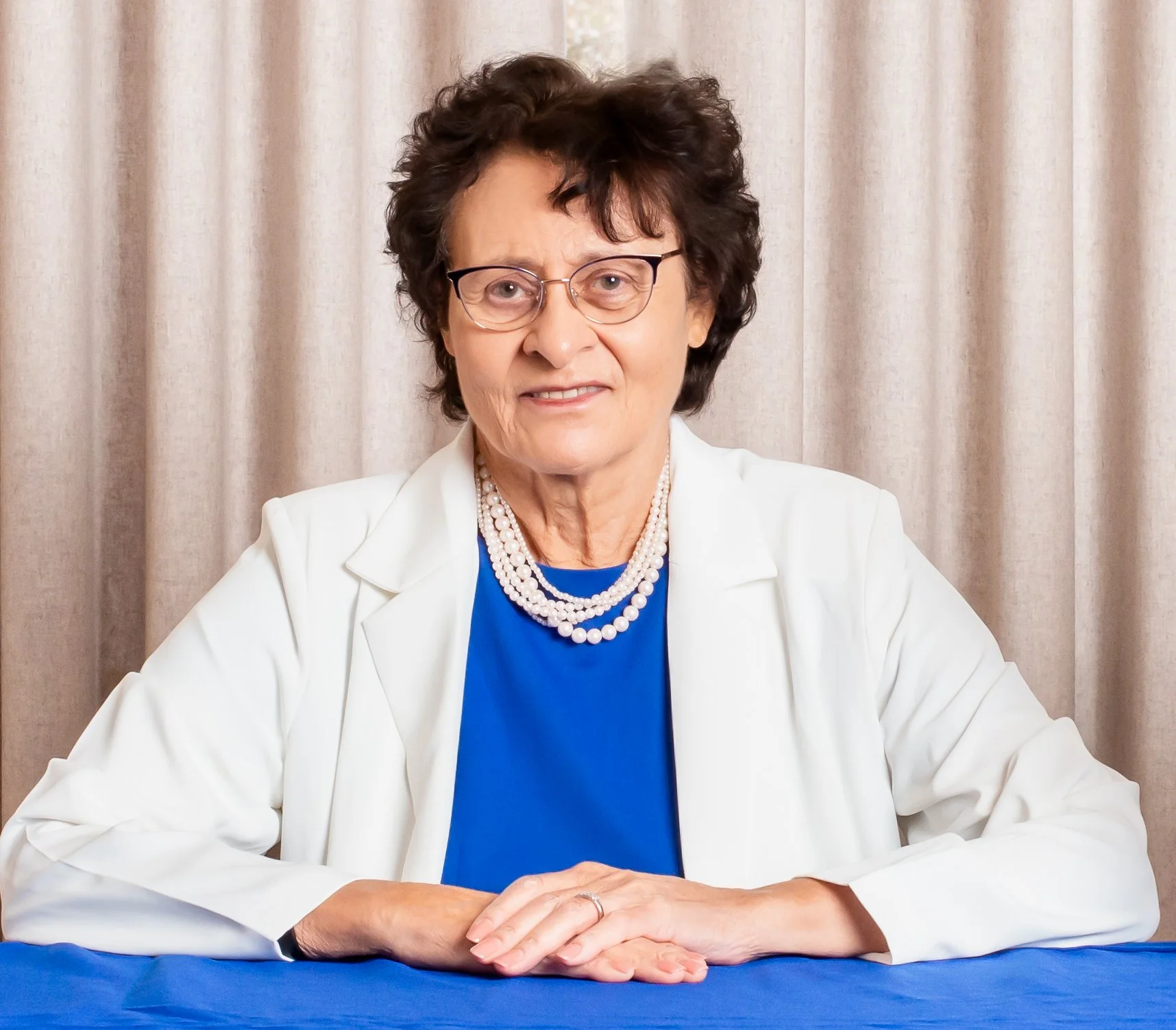 An elderly woman wearing glasses, a white blazer, a blue top, and pearl necklaces, sitting at a table with hands clasped, in front of a beige curtain.