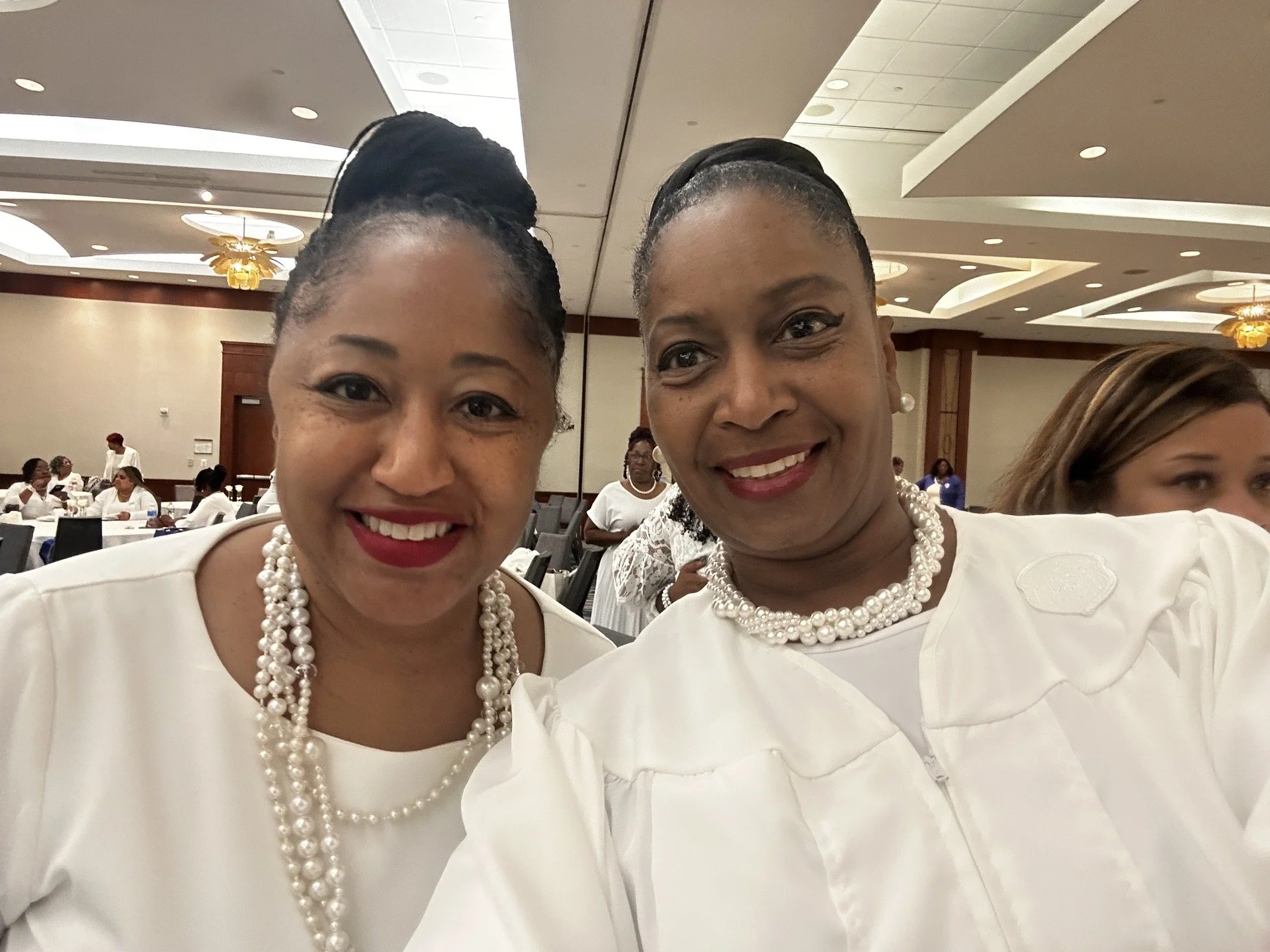 Two women dressed in white, wearing pearl necklaces, taking a selfie at a formal event with many other attendees seated at round tables in the background.