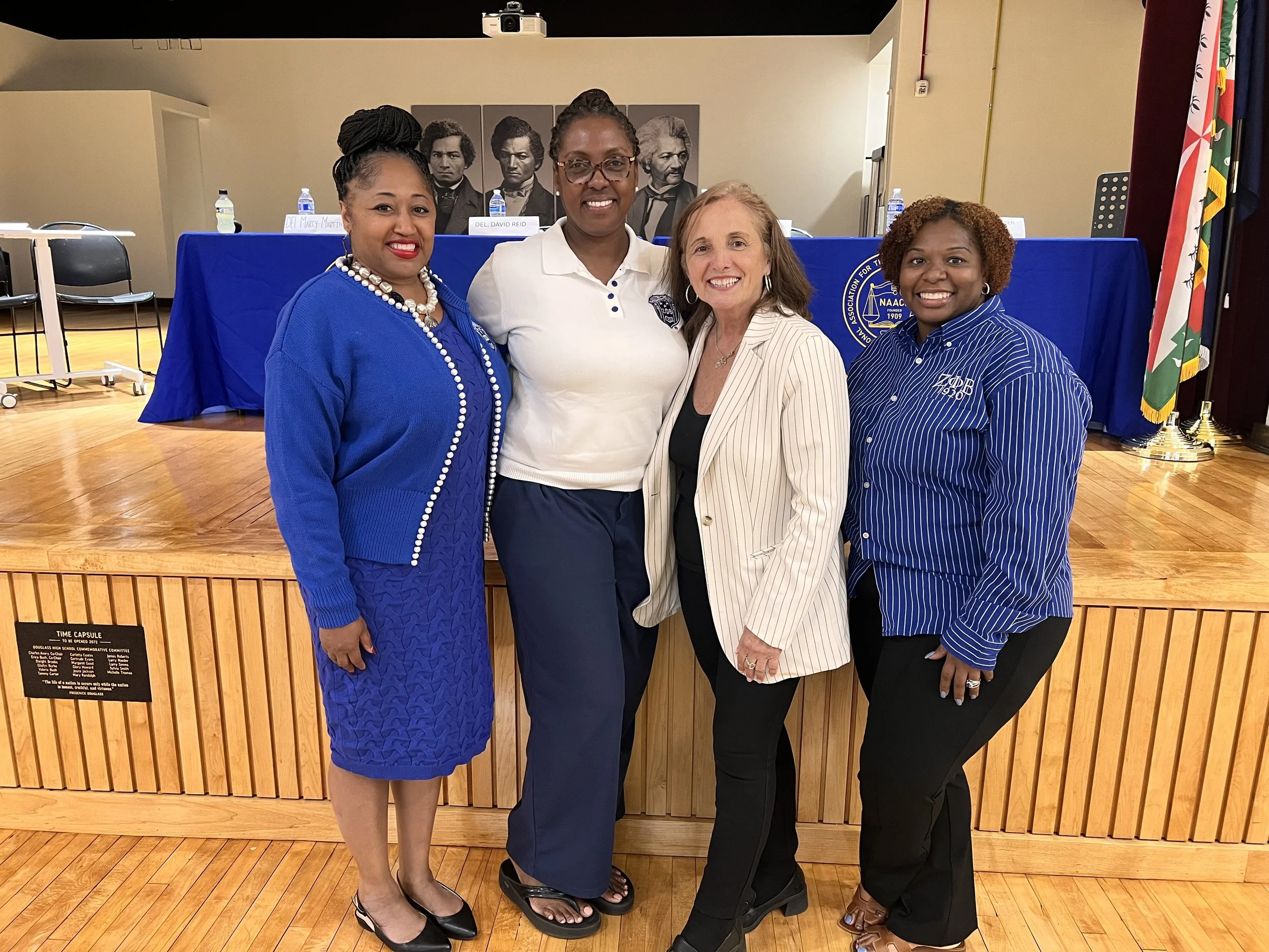 Four women standing together in front of a blue table with water bottles and nameplates, smiling for the photo at an indoor event.