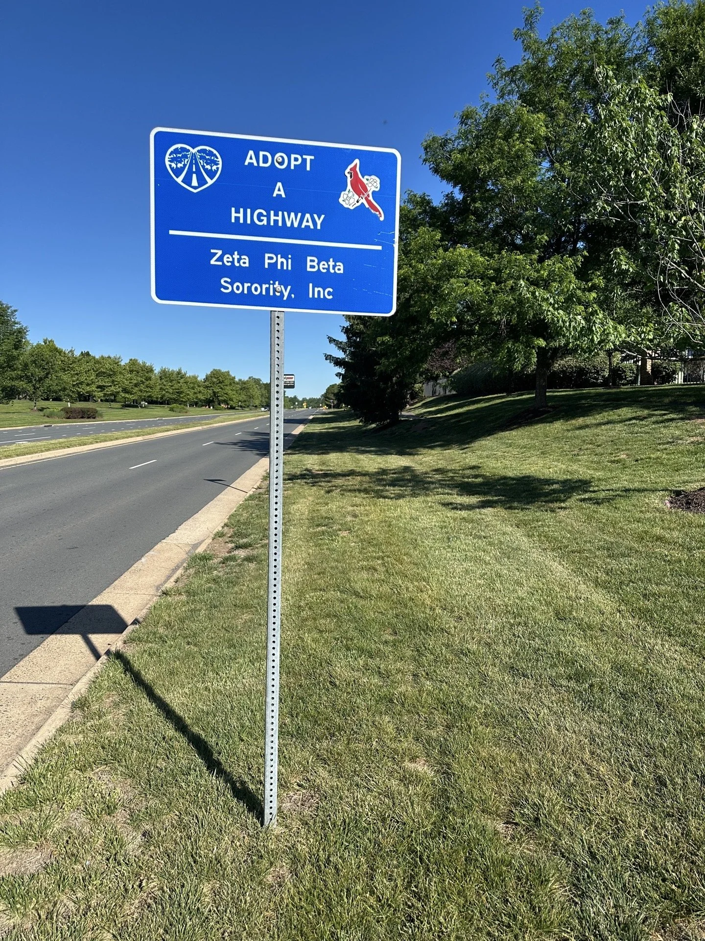Blue highway sign encouraging adoption of a highway, with symbols of a heart with a road and trees, and a red cardinal bird sticker.