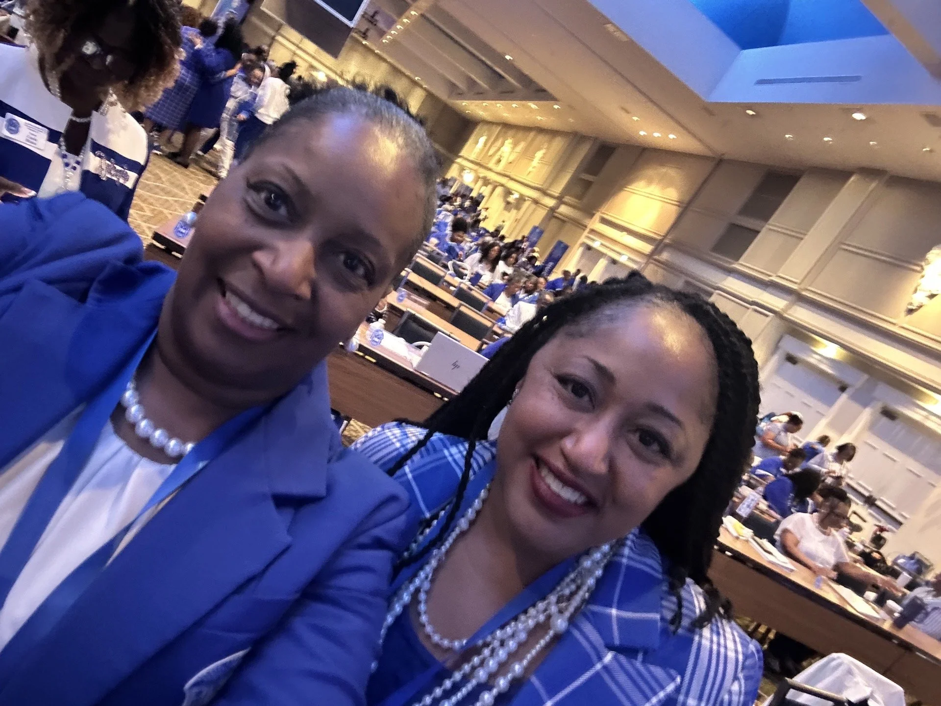 Two women smiling at the camera, attending a conference or large meeting in a spacious, well-lit room filled with many people seated at tables, many wearing blue shirts.