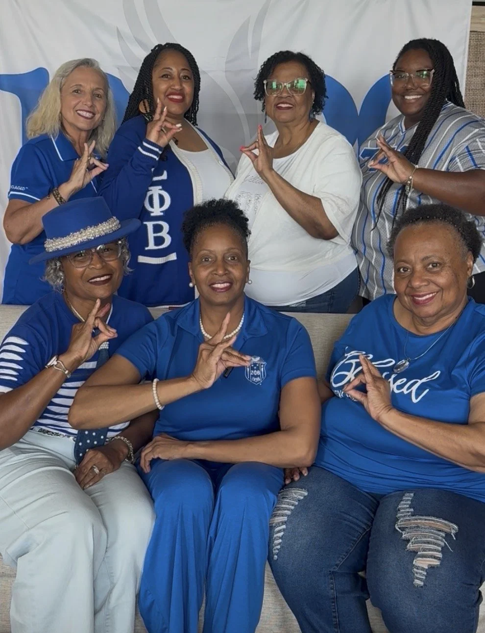 Group of women posing together, with some making the 'OK' sign with their hands, at a celebration or event, wearing blue and white clothing.