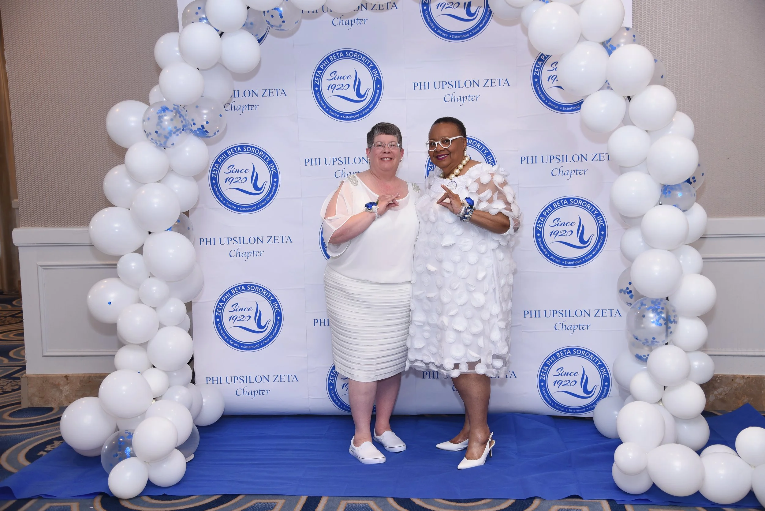 Two women standing in front of a backdrop with the logo of Phi Upsilon Zeta Sorority and balloons framing the scene. One woman is wearing a white outfit with sheer details, and the other is wearing a white dress with circular patterns. Both are smili