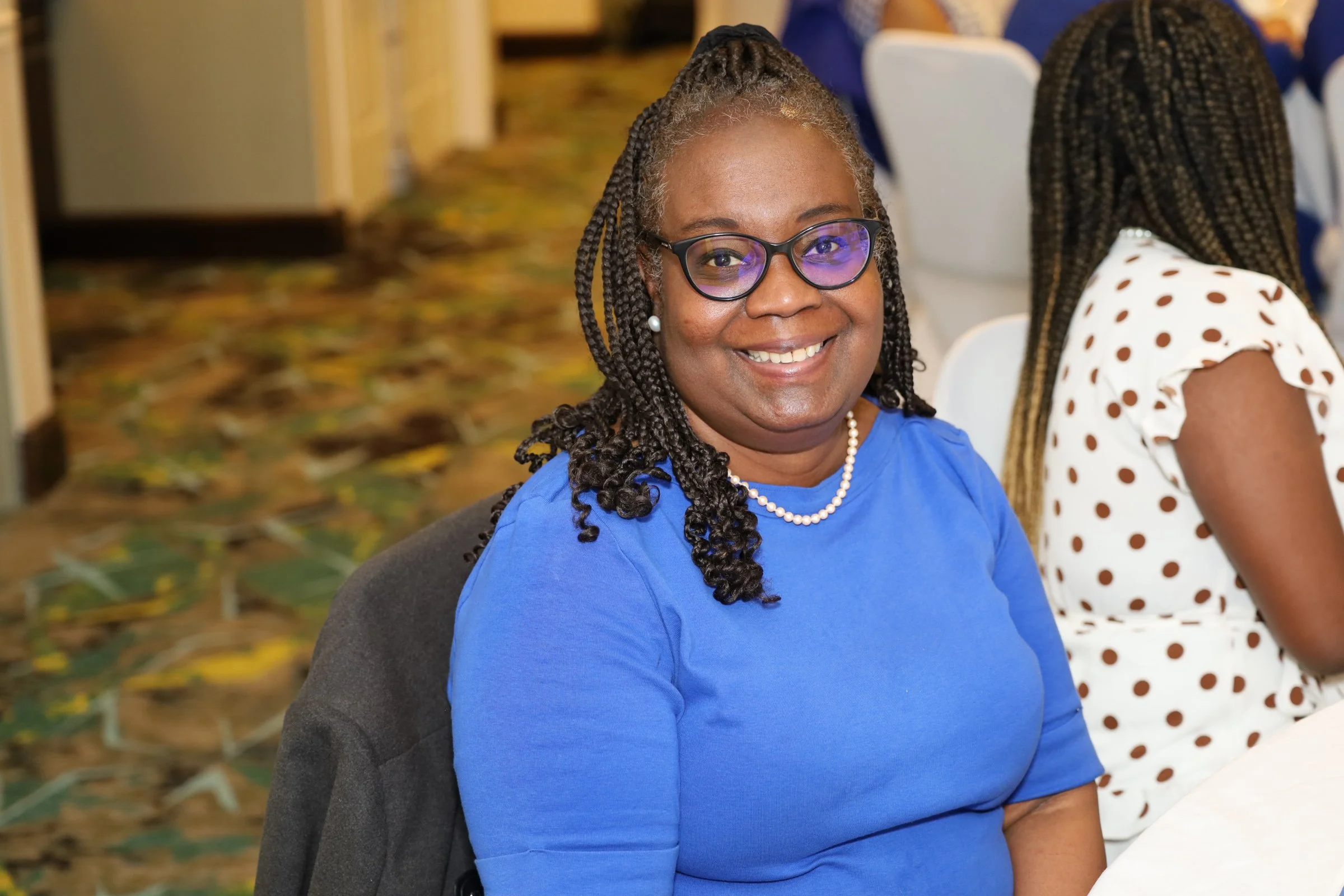 A smiling African American woman wearing glasses, a blue shirt, and a pearl necklace sitting at a conference or meeting table.