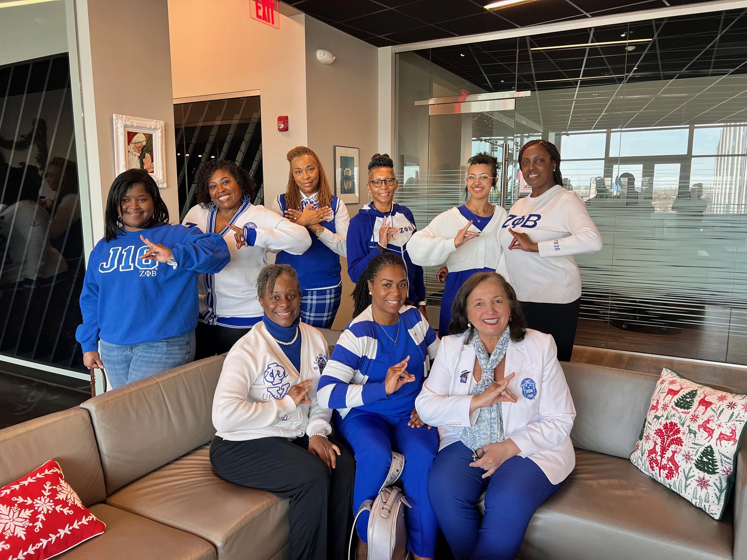 A group of women, some wearing blue and white coordinated outfits with Greek letters, posing for a photo in a modern indoor space with glass walls and framed artwork. Some women are standing, others sitting, and they are making positive hand gestures