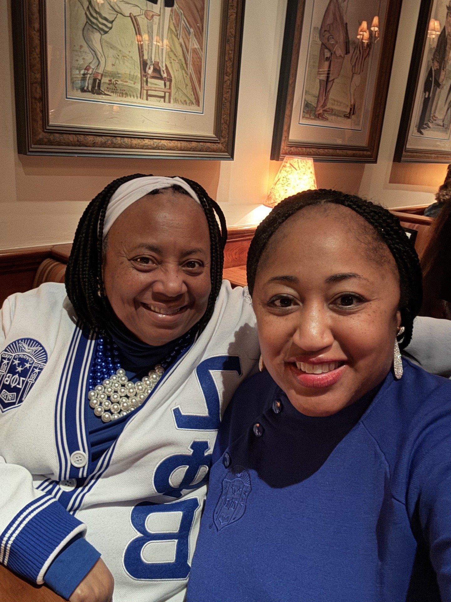 Two women smiling and taking a selfie, seated in a cozy restaurant with framed artworks and warm lighting in the background.