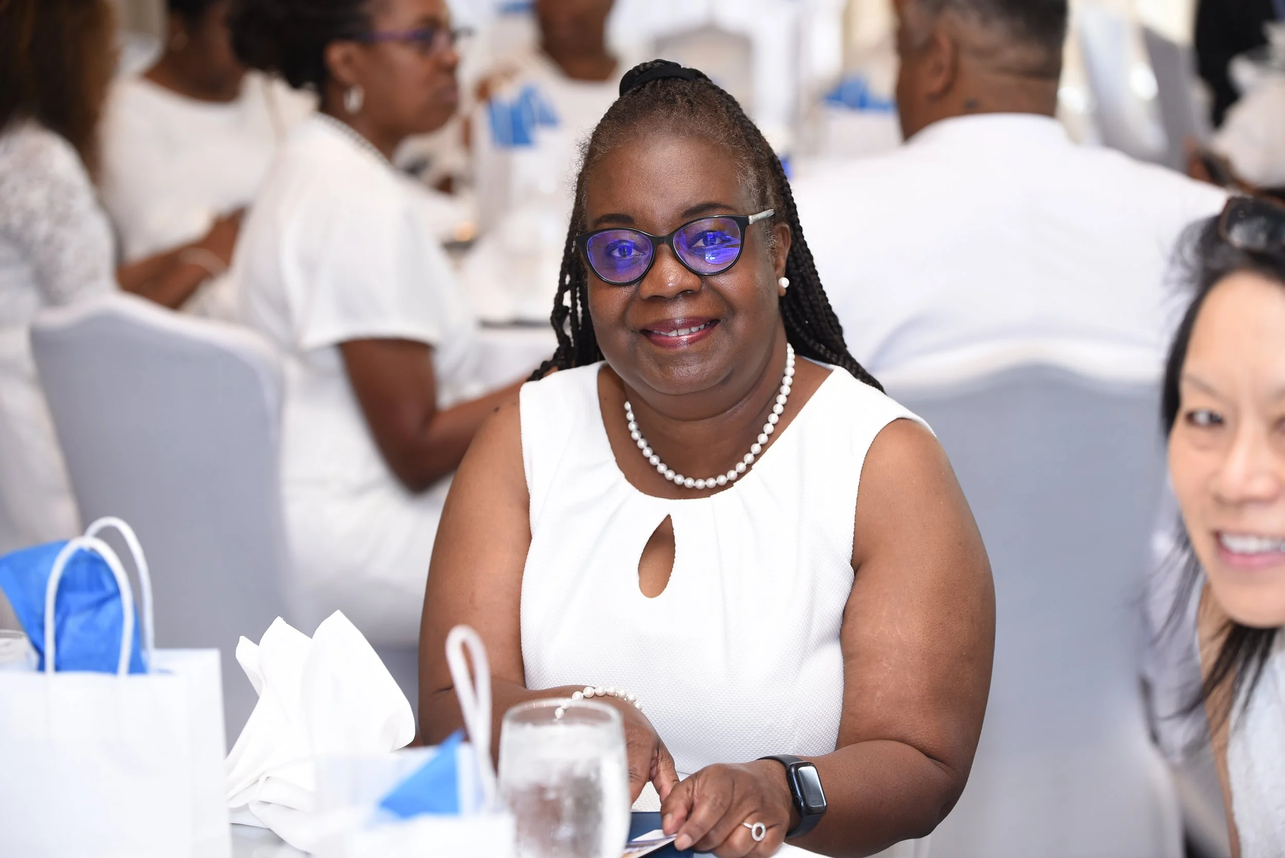 Smiling woman with glasses, braided hair, pearl necklace, and white sleeveless top sitting at a table during a social event.