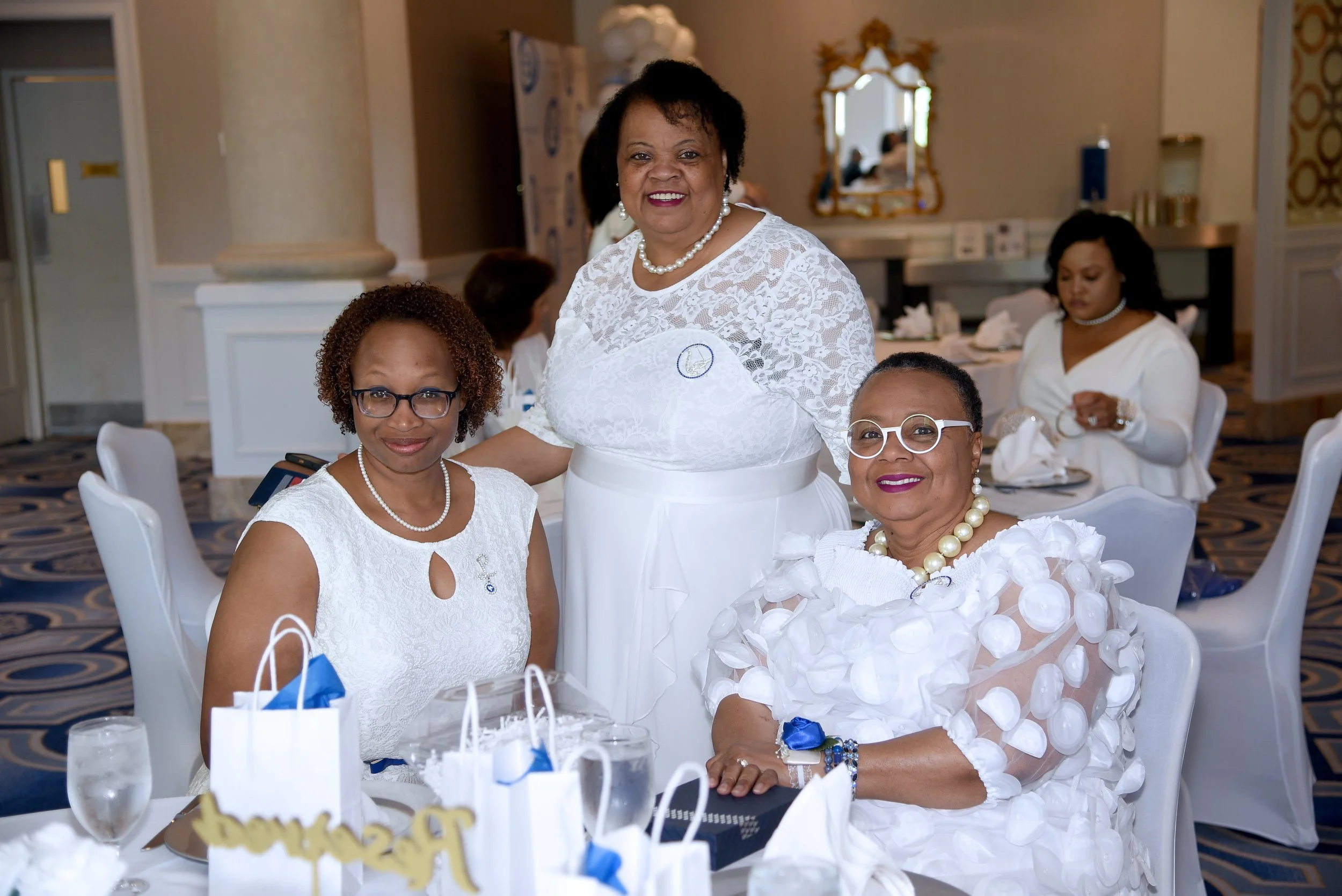 Four women dressed in white at a formal event, with a woman standing and three seated around a table with gifts and decorations. They are smiling and wearing pearl jewelry.