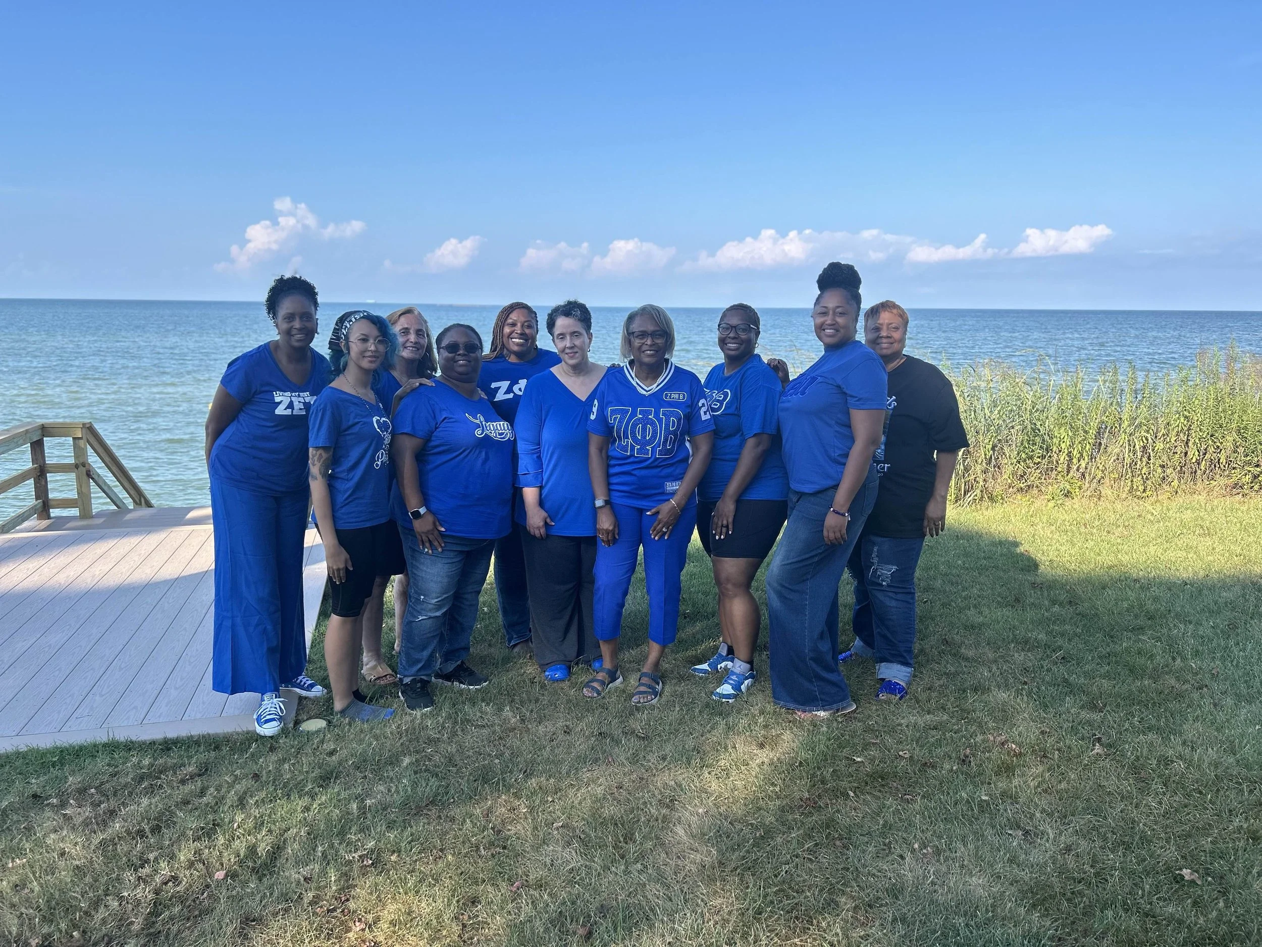 A group of ten women standing together on a grassy area near the water, with a wooden deck on the left and a clear blue sky with some clouds in the background. Most women are wearing blue shirts, some with Greek letters on them, and all are smiling.