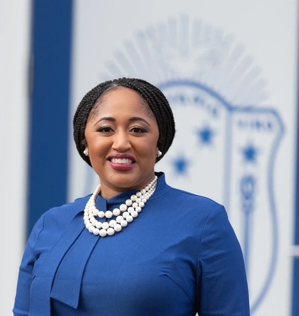 A smiling woman wearing a blue blazer and white pearl necklace, standing in front of a blurred police badge backdrop.
