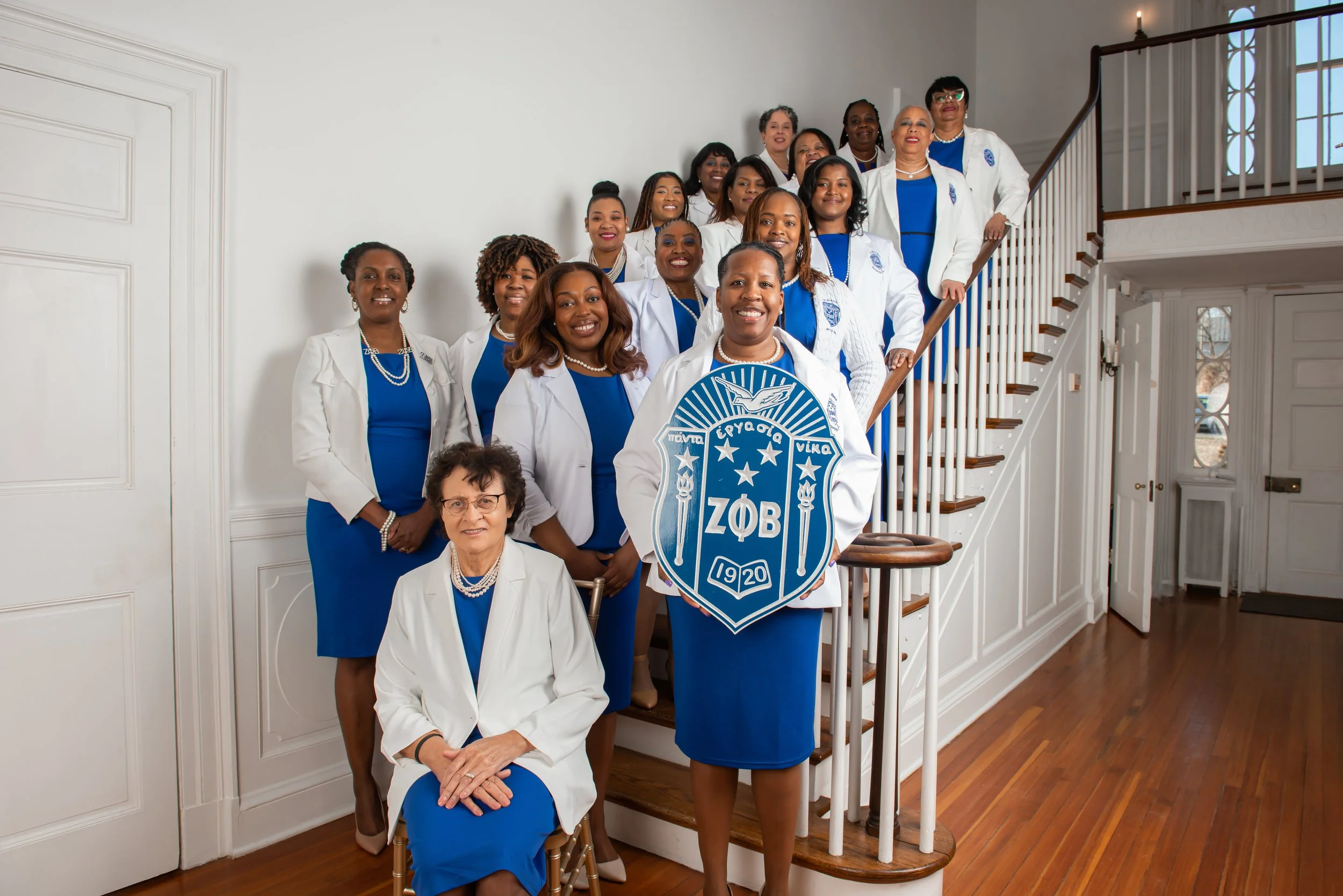 A group of women dressed in blue dresses and white blazers, with some holding a fraternity shield, standing on a staircase inside a house with white walls and wooden flooring.
