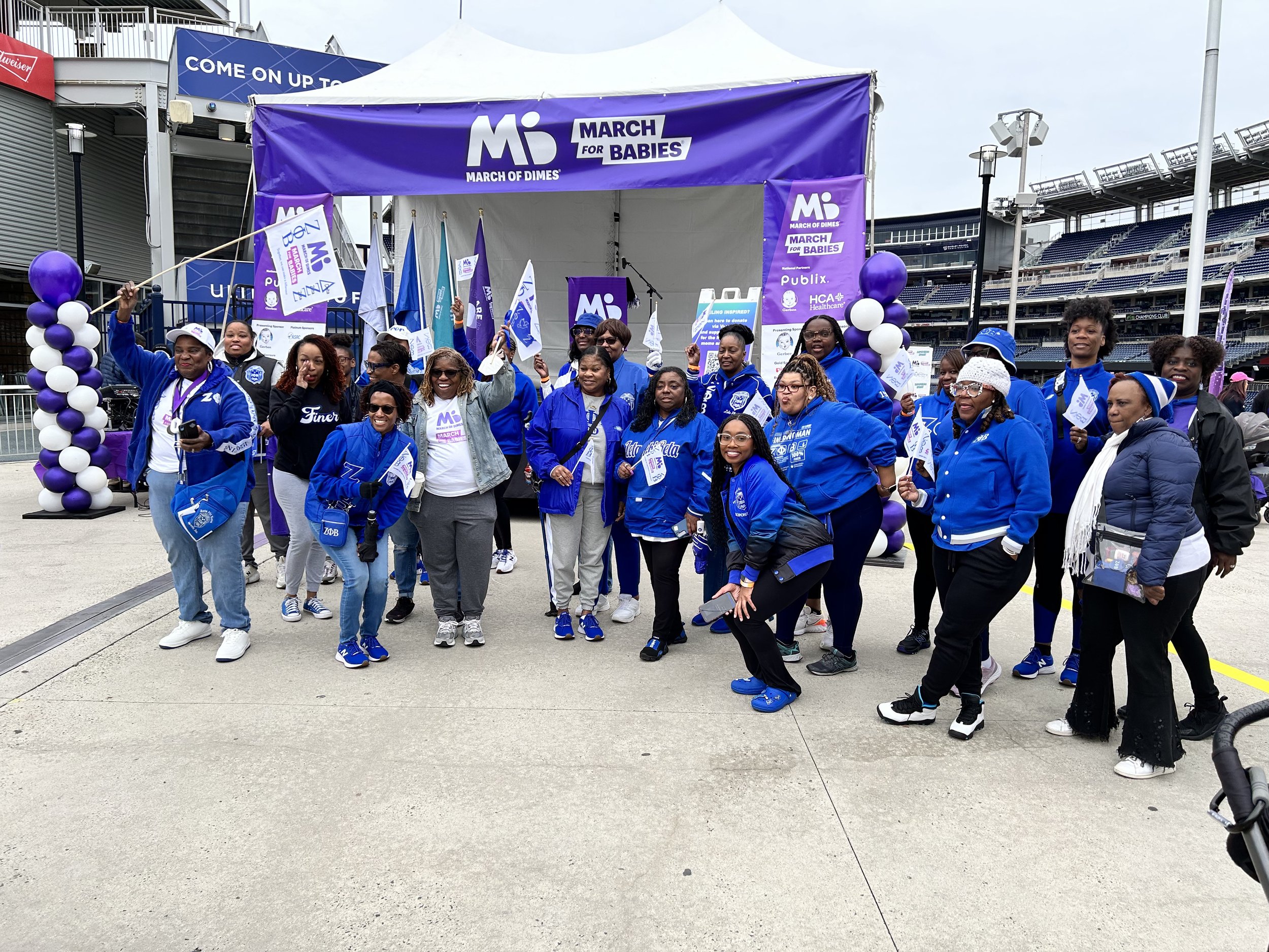 A group of diverse women and men gather in front of a stage with purple banners and balloons, participating in the March for Babies event at a stadium.