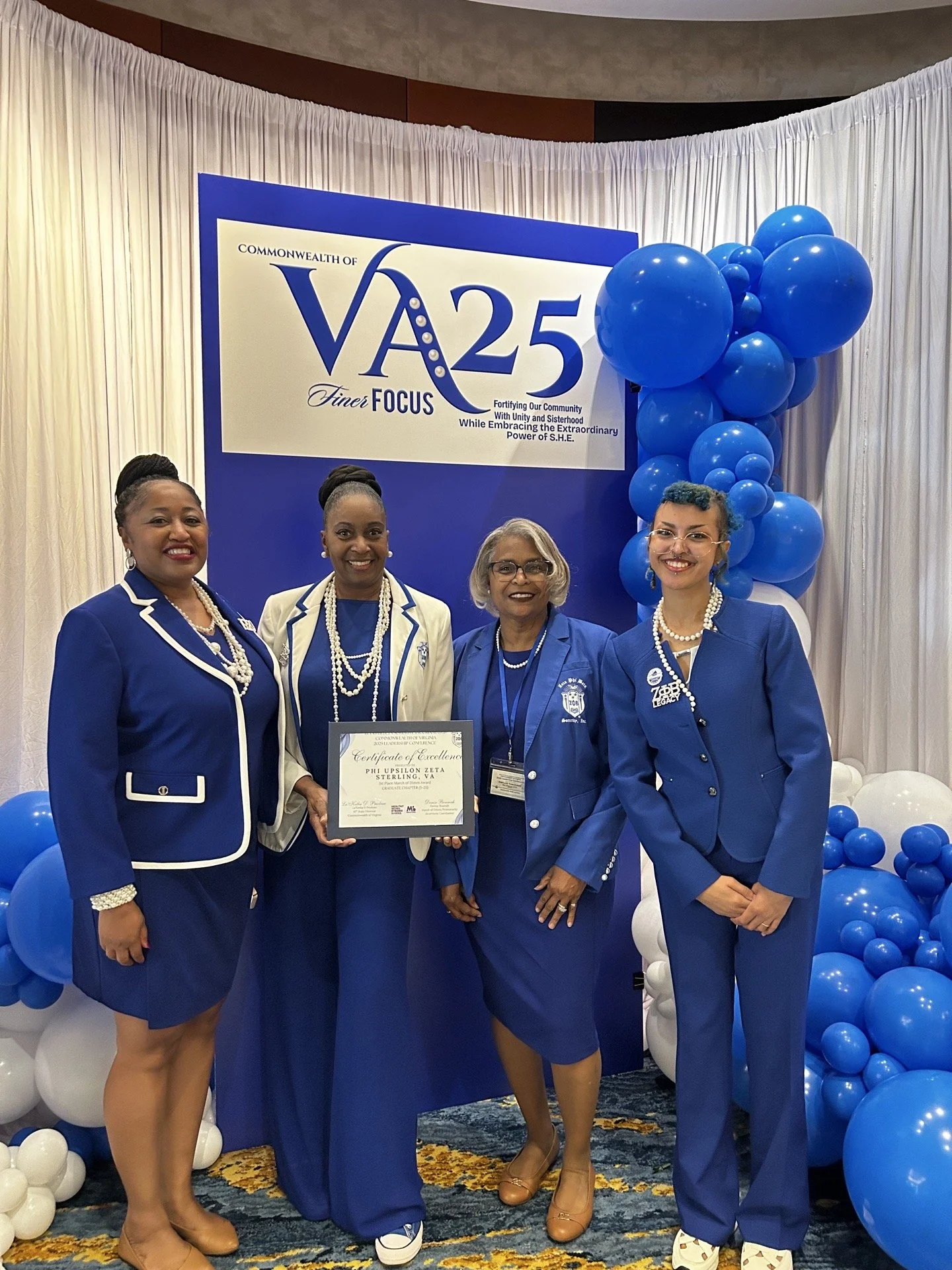 Four women standing together in front of a large blue and white banner for an event, with blue and white balloon decorations. One woman is holding a certificate. The women are dressed in blue and white business attire.