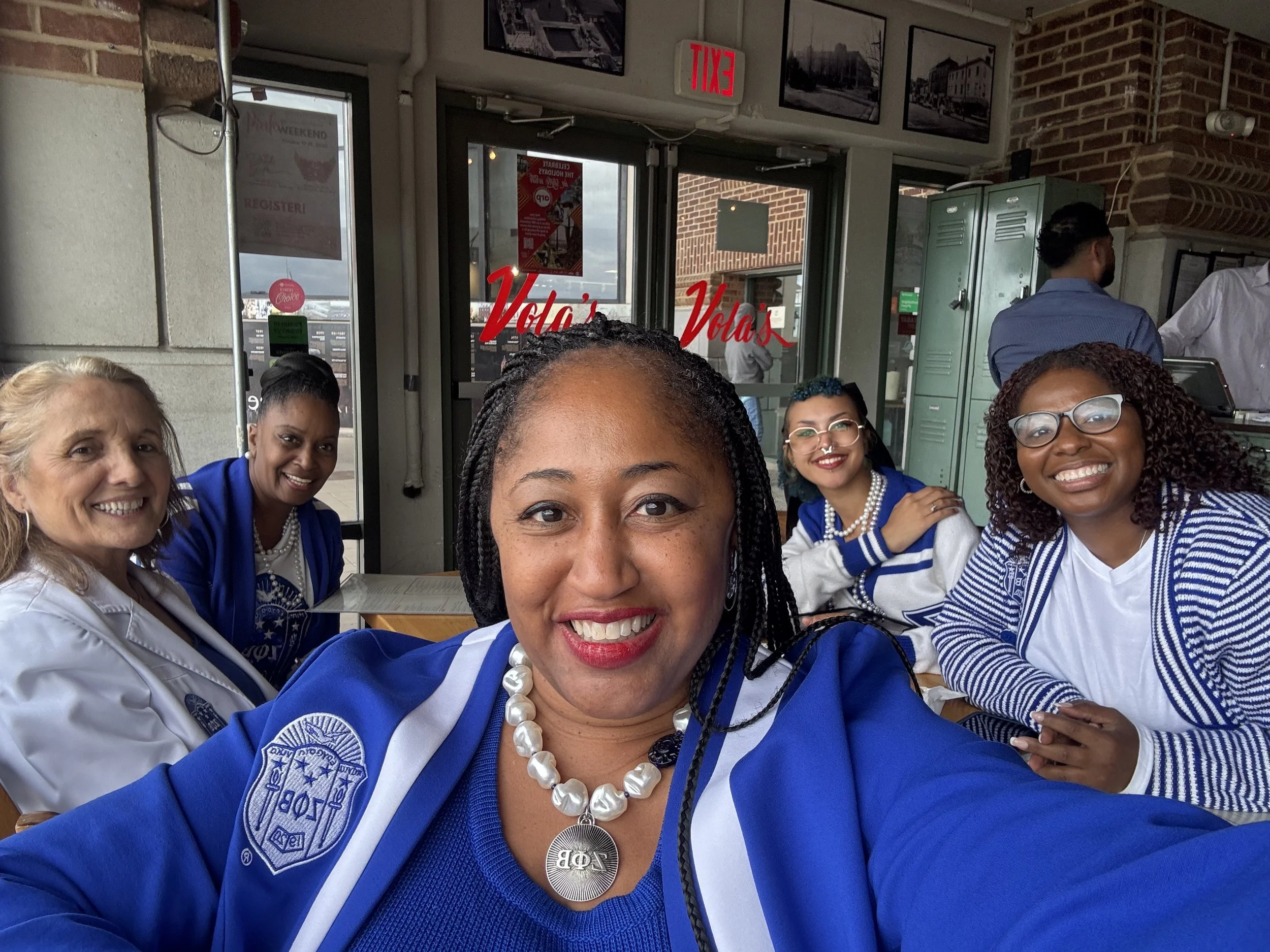 A group of diverse women smiling and taking a selfie at a restaurant. The women are wearing blue and white uniforms with embroidered patches, and necklaces. The background shows a window with the words 'Vola' and 'Vola's' written in red, and a door w