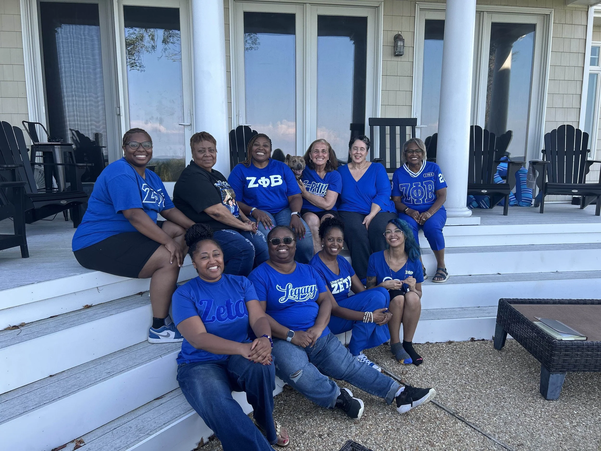 Group of women and a dog sitting and standing on the steps of a house, all wearing blue shirts, with a glass door and black chairs behind them.