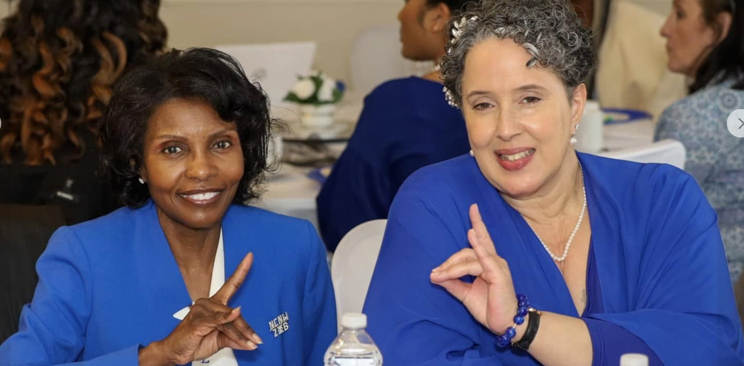 Two women in blue suits sitting at a table, making a heart shape with their hands, smiling at the camera. There are other women in the background.