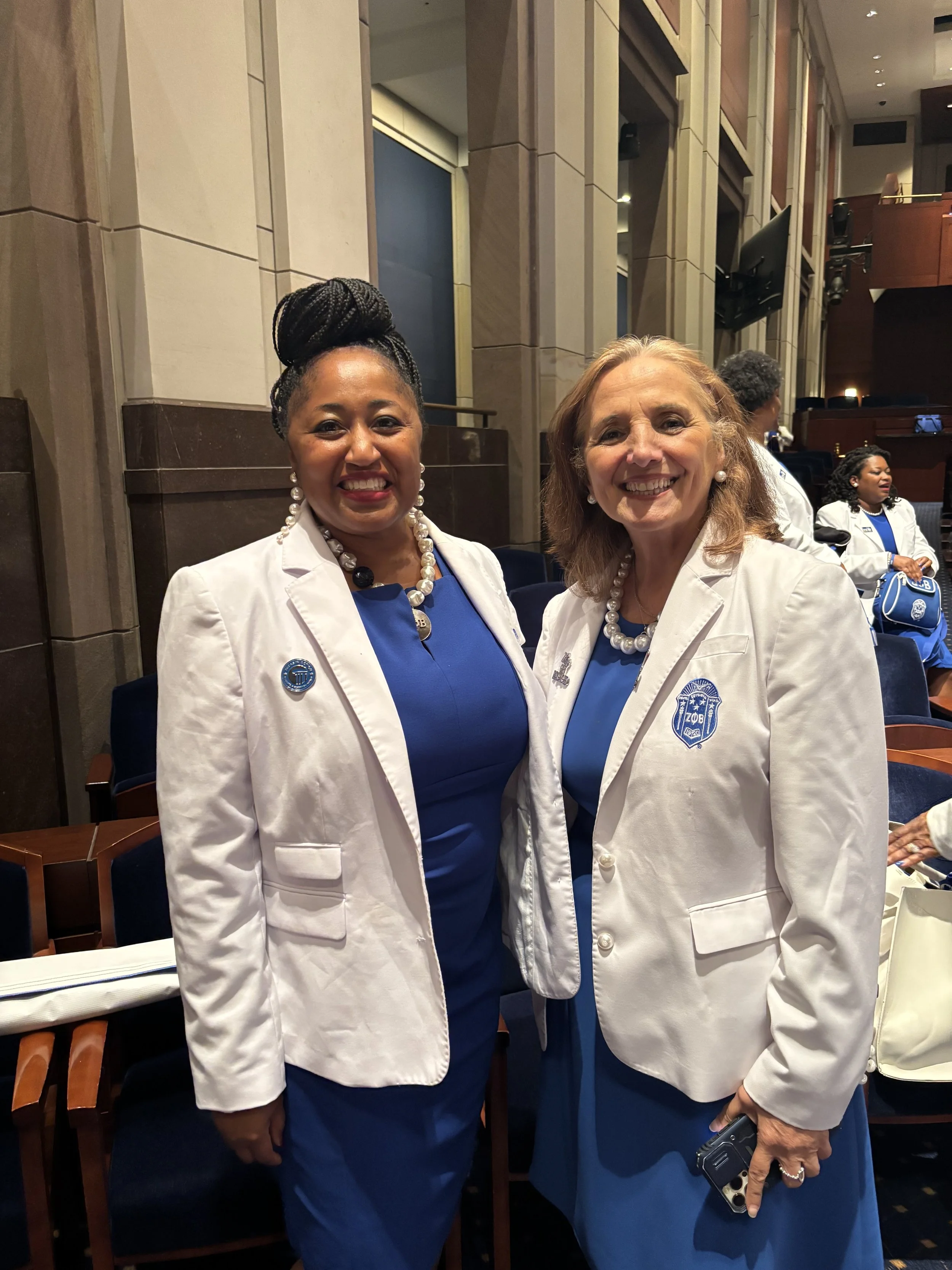 Two women dressed in white blazers with blue dresses underneath, wearing pearl necklaces and earrings, standing indoors at a formal event.
