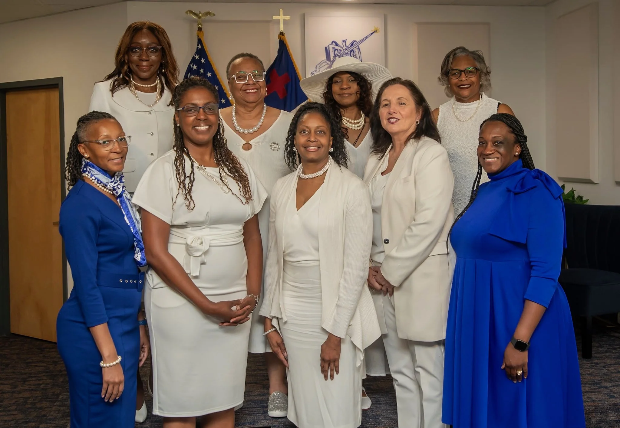 A group of eleven women standing indoors in front of two flags and a religious cross, dressed in white and blue clothing, smiling at the camera.