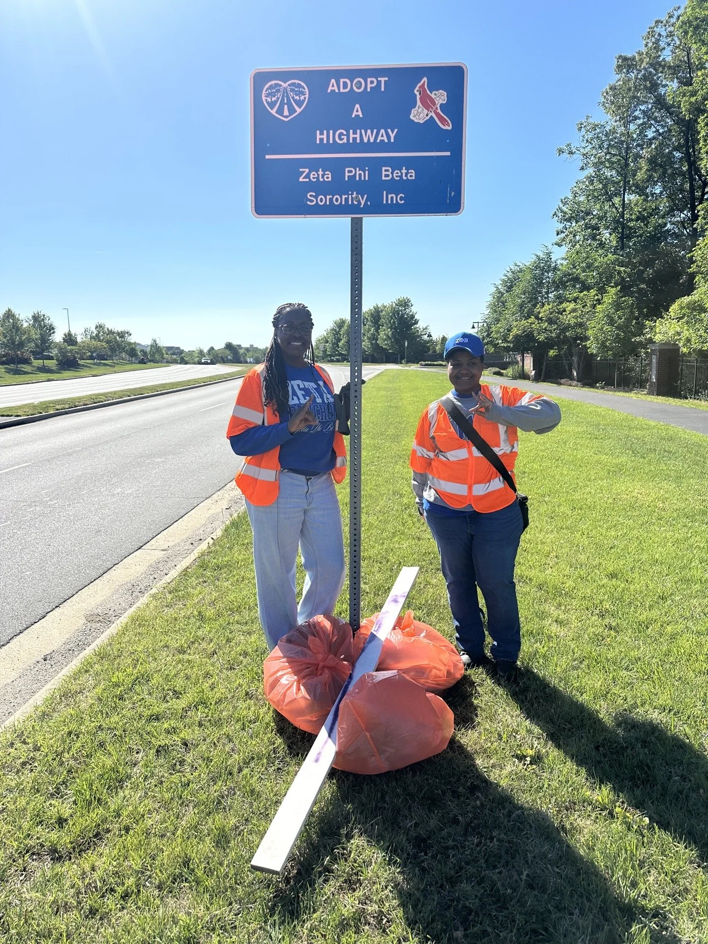 Two women in orange safety vests posing next to a highway sign for adopting a highway, with trash bags and a long white stick on the grass.