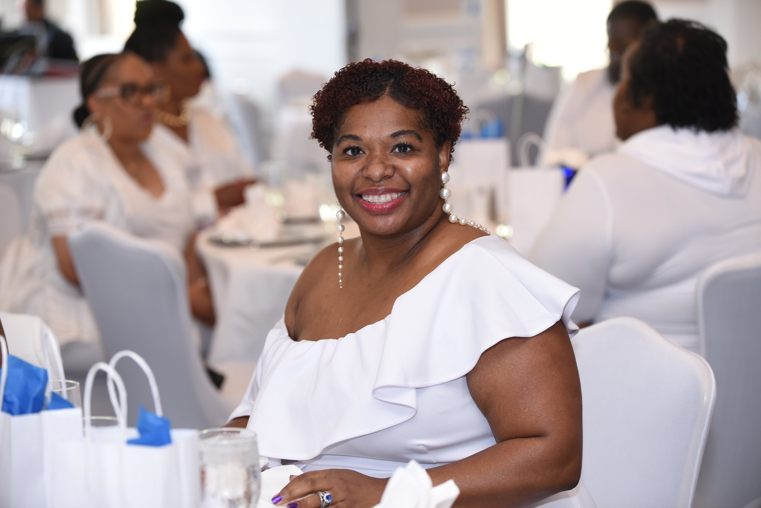 A woman with short curly hair and pearl earrings smiling at the camera during a formal event with other women in the background dining at tables.