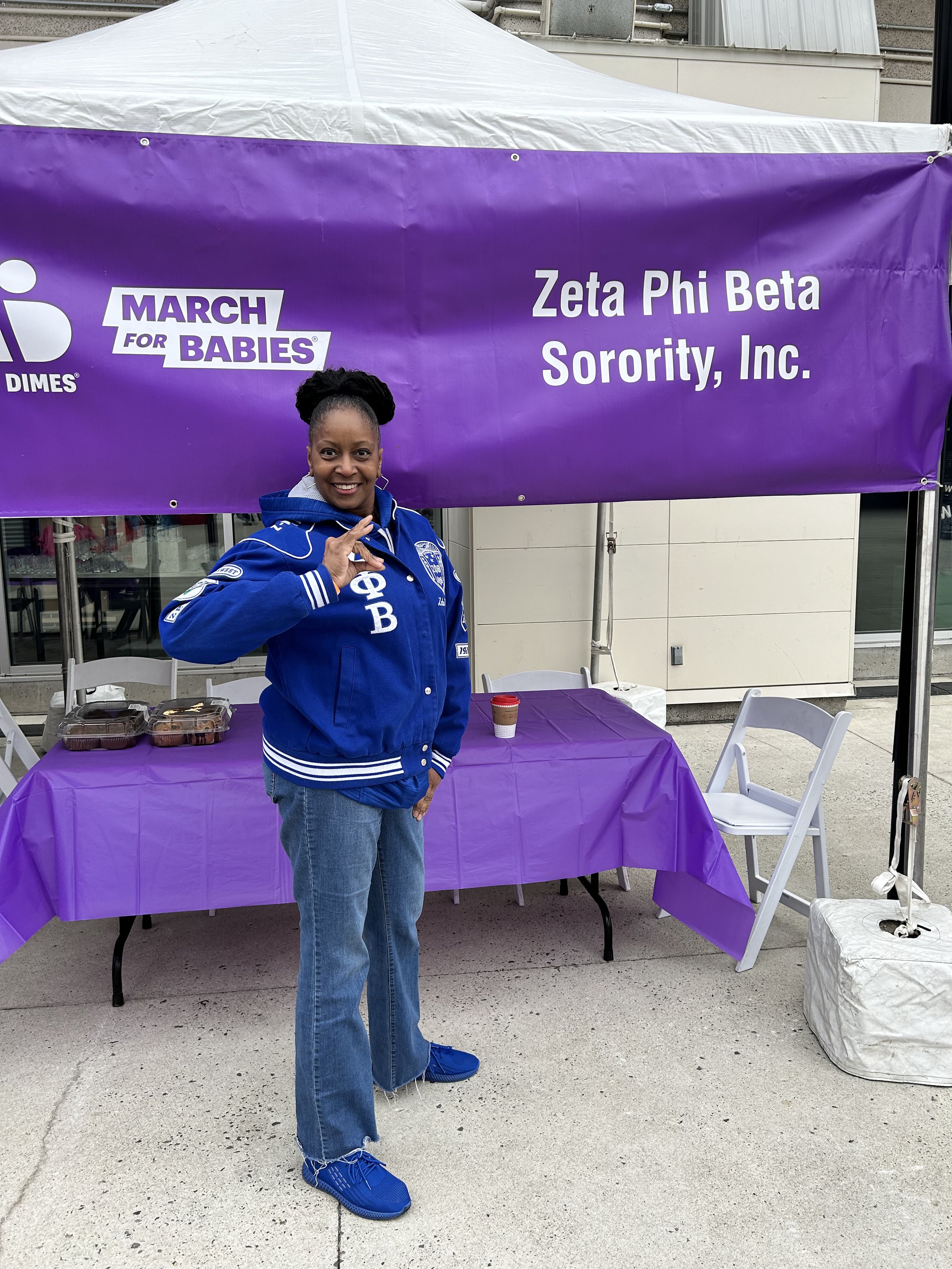 A woman standing in front of a purple table with food containers, making an okay hand gesture, at an outdoor event under a purple and white tent with a large banner that reads 'March for Babies' and 'Zeta Phi Beta Sorority, Inc.'