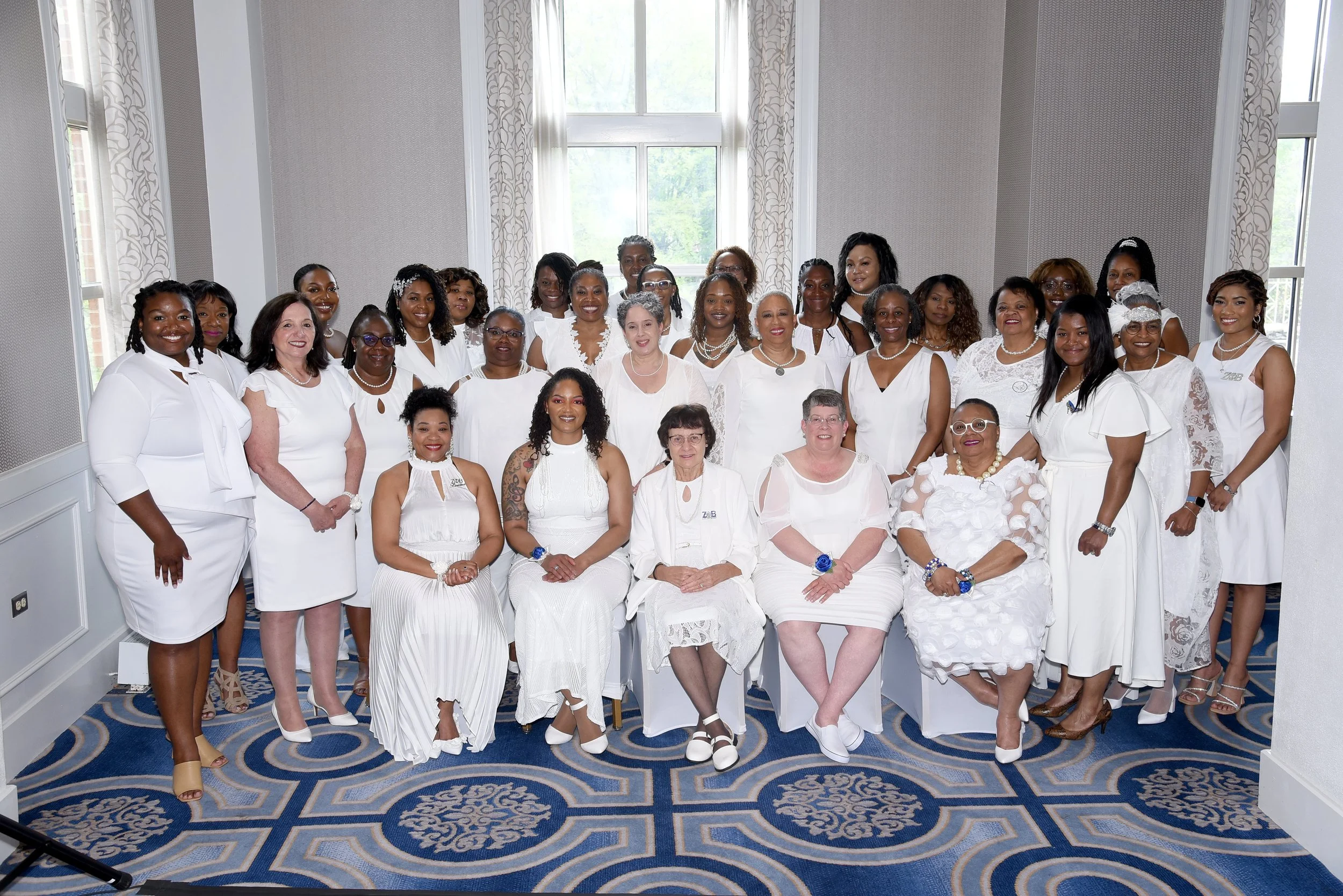 Group of women dressed in white, smiling, gathered in a brightly lit room with large windows and decorative curtains.