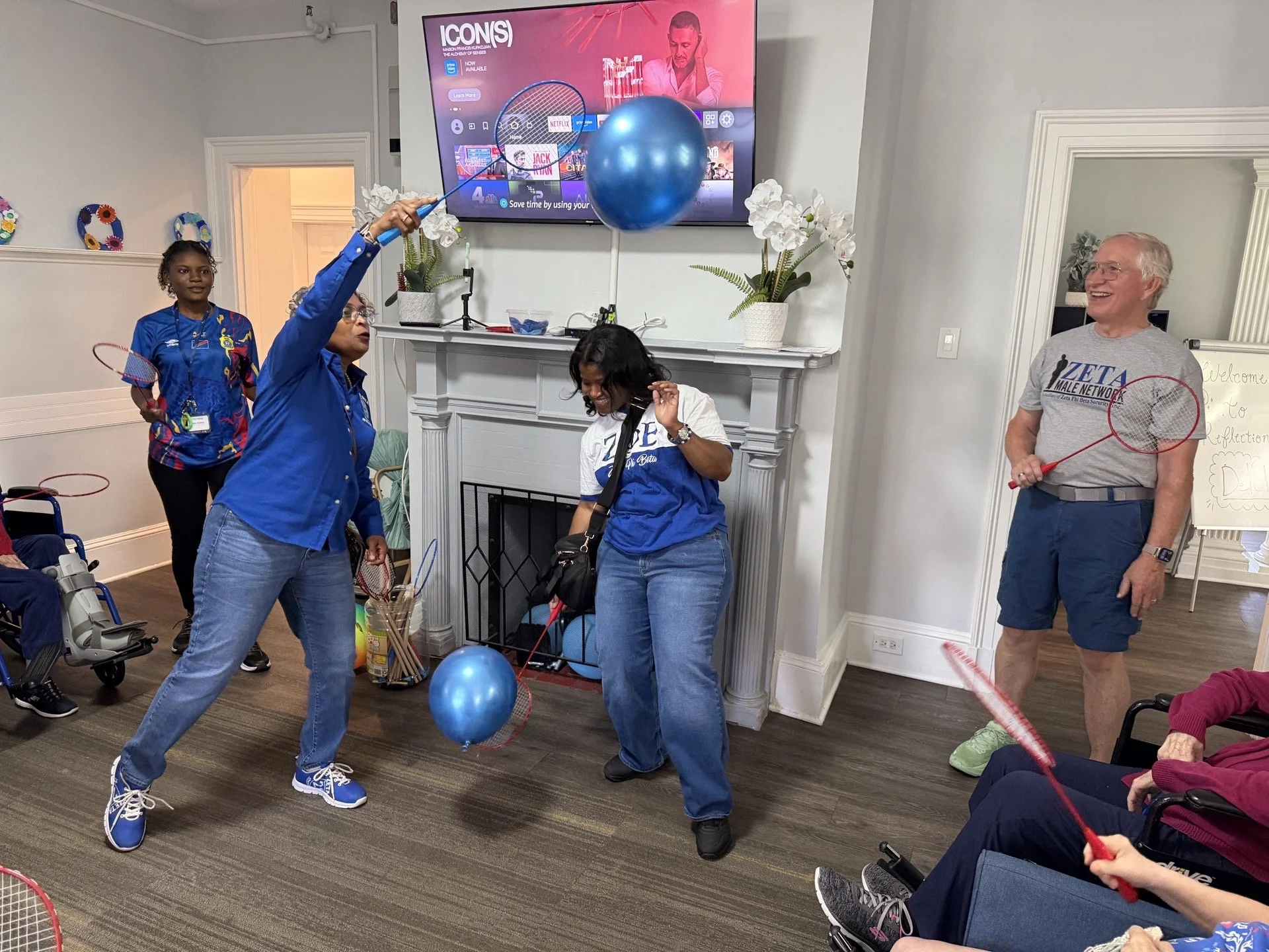 Several people are playing a game with balloons and small rackets indoors. A woman in a blue jacket and jeans is hitting a balloon, while another woman in a white t-shirt is observing and a man with glasses and a gray t-shirt is smiling, holding a ra