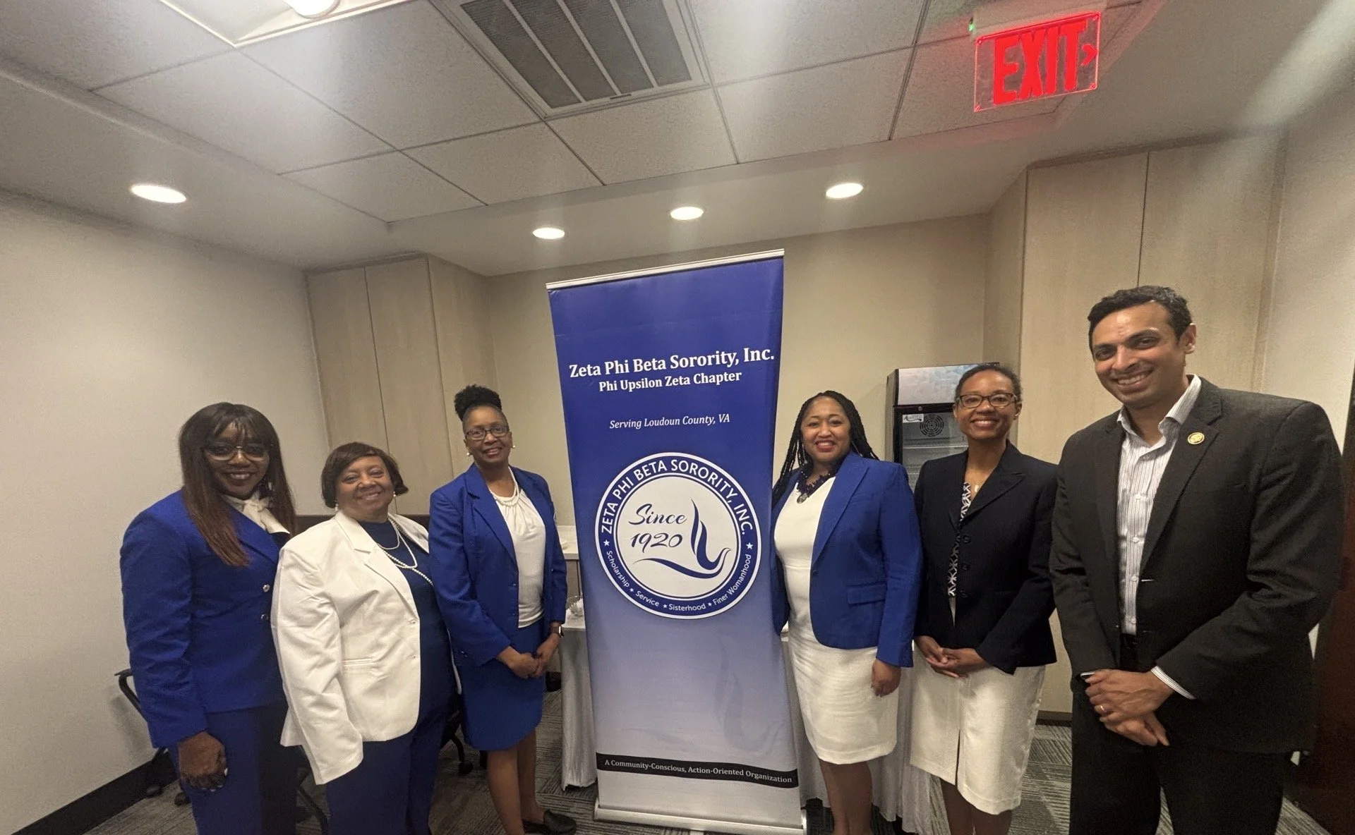 Group of six professionally dressed women and one man standing together at an event, posing in front of a blue banner that reads 'Zeta Phi Beta Sorority, Inc. Phi Upsilon Zeta Chapter,' with the organization logo and details.