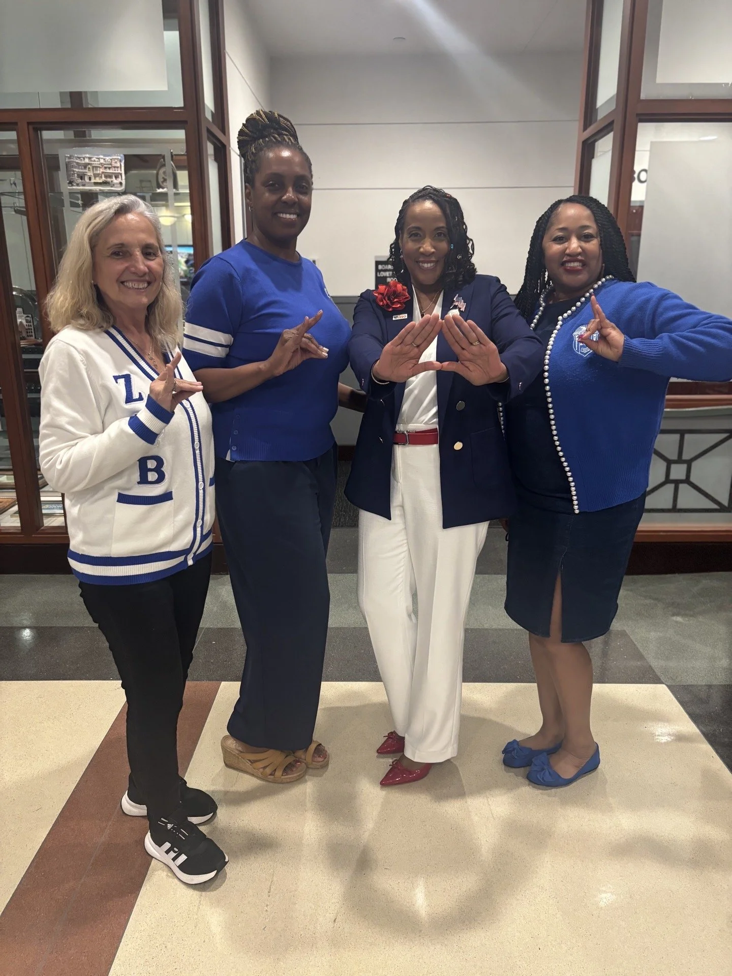 Four women standing and smiling, making hand gestures, dressed in blue and white clothing, in an indoor setting with a tiled floor and wood-paneled display cases.