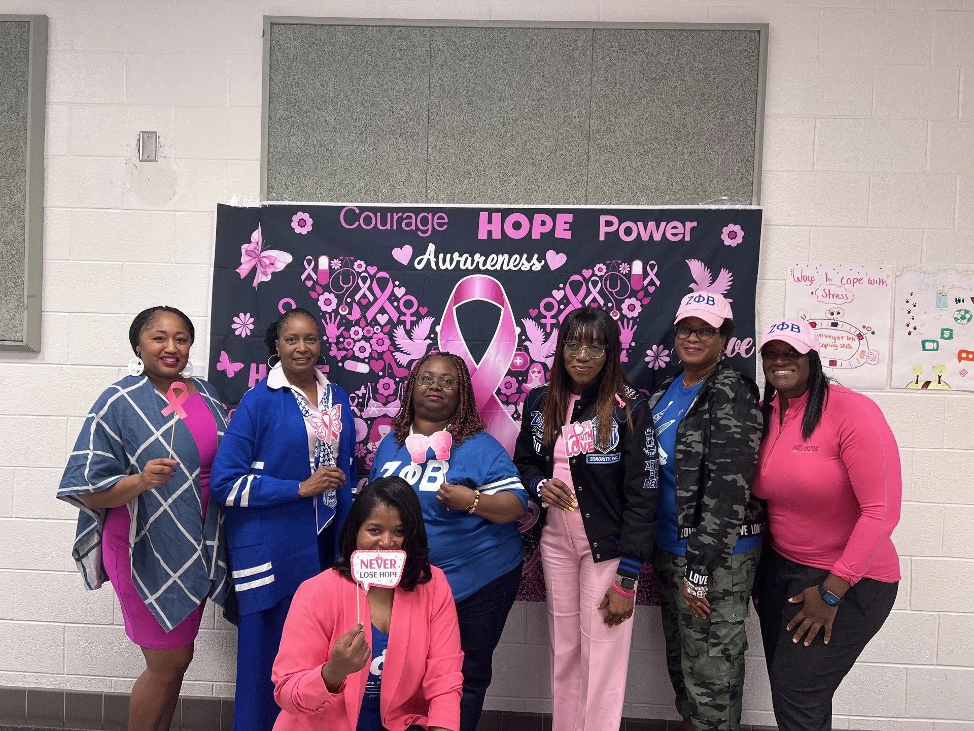 Group of women standing together in front of a pink awareness banner related to breast cancer, with some holding pink ribbons and signs supporting hope and love.