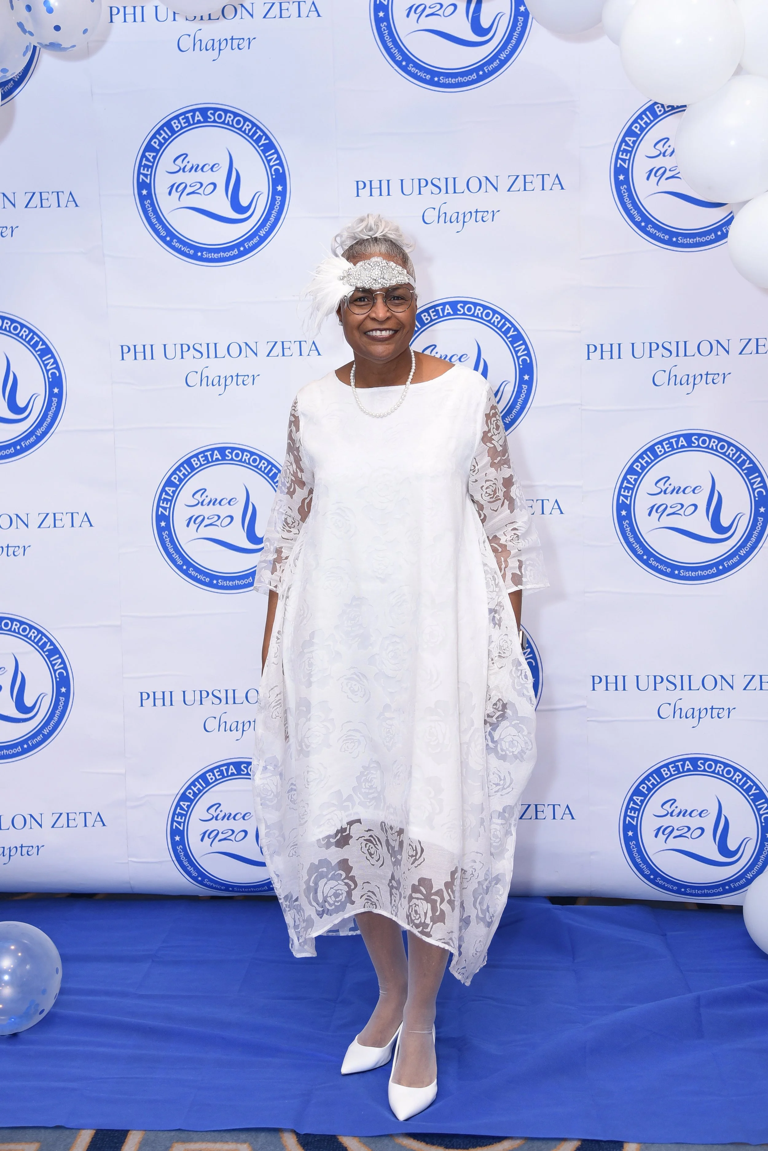A woman dressed in a white lace dress with floral patterns, wearing white heels, pearl necklace, and a decorative headpiece with feathers, standing in front of a backdrop with logos and text for Phi Upsilon Zeta chapter, surrounded by white balloons.