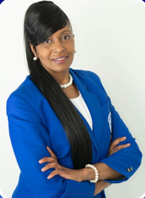 A woman with long black hair, wearing a blue blazer, white blouse, pearl necklace, and pearl earrings, standing with arms crossed against a white background.