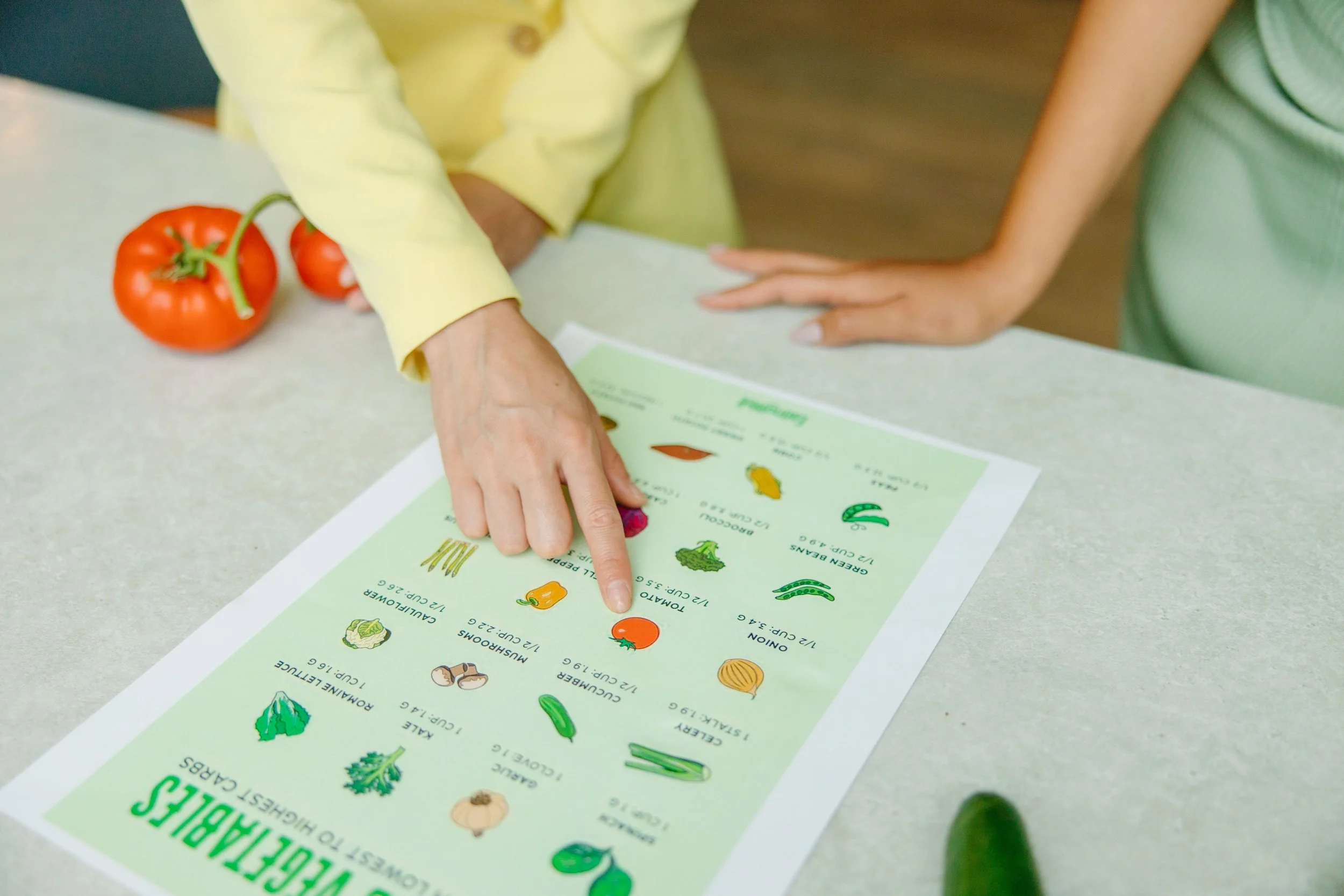 Deux personnes regardent et pointent un affichage éducatif sur les légumes, avec des illustrations et descriptions, sur une table, accompagnés de tomates rouges.