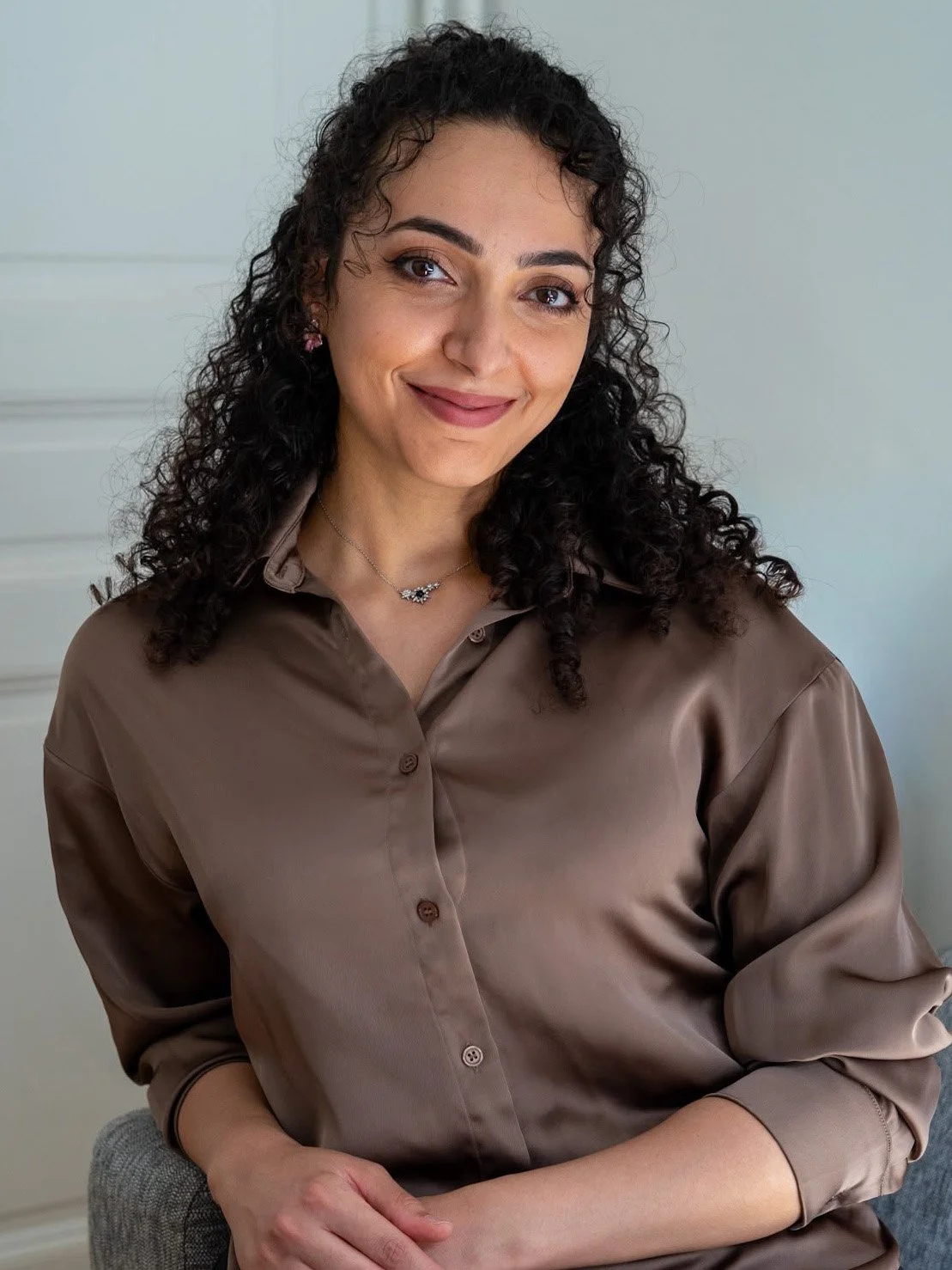 Une femme souriante avec des cheveux bouclés portant une chemise brune, portant un collier et des boucles d'oreilles, assise dans un intérieur clair.