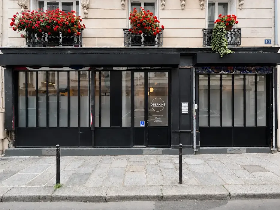 Façade de restaurant ou café avec fenêtres à carreaux noirs, porte d'entrée centrale, et balcons avec fleurs rouges au-dessus. En bas, trottoir pavé et barrières pour piétons.