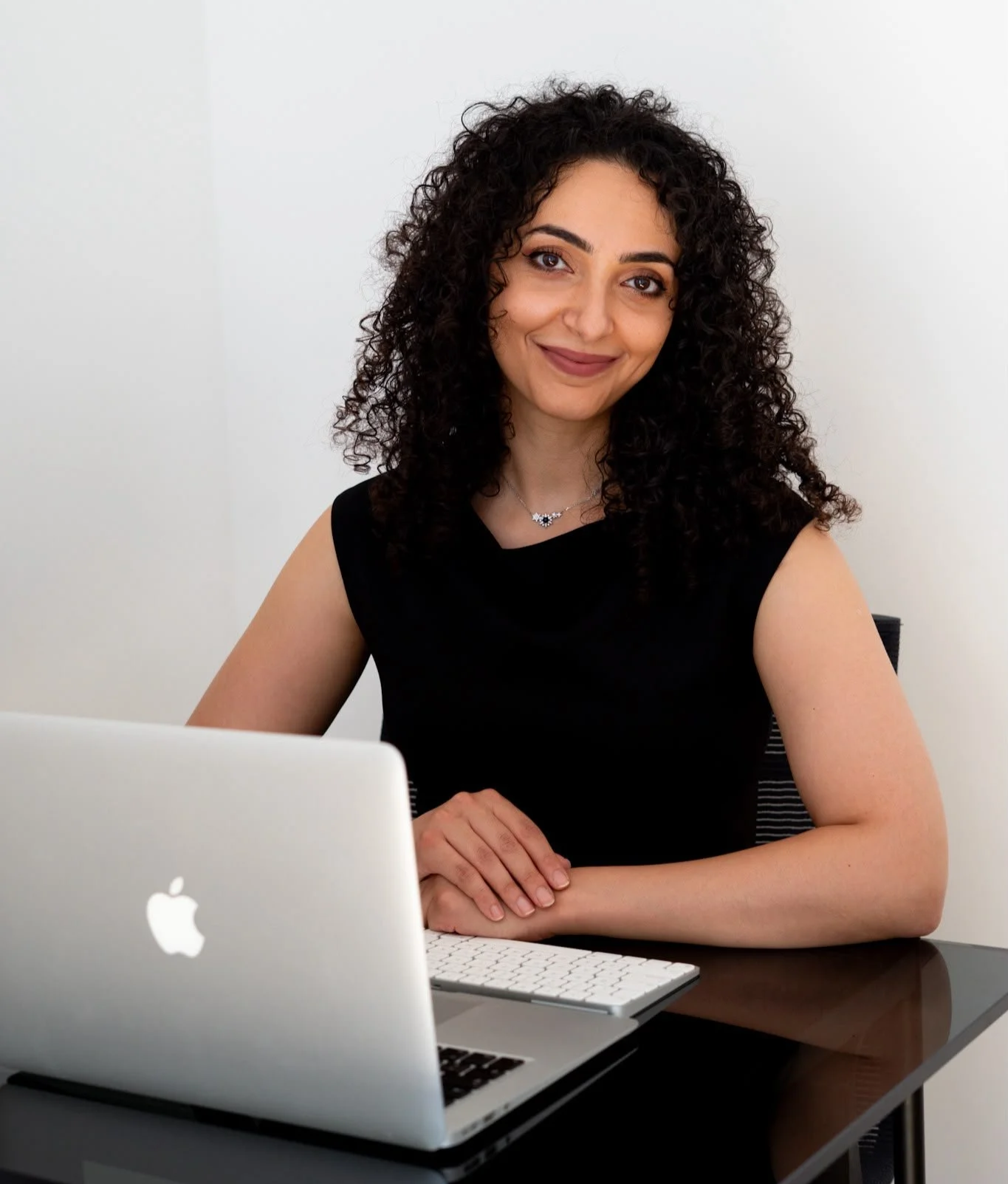 Une femme souriante avec des cheveux bouclés assise devant un ordinateur portable blanc dans un bureau blanc.