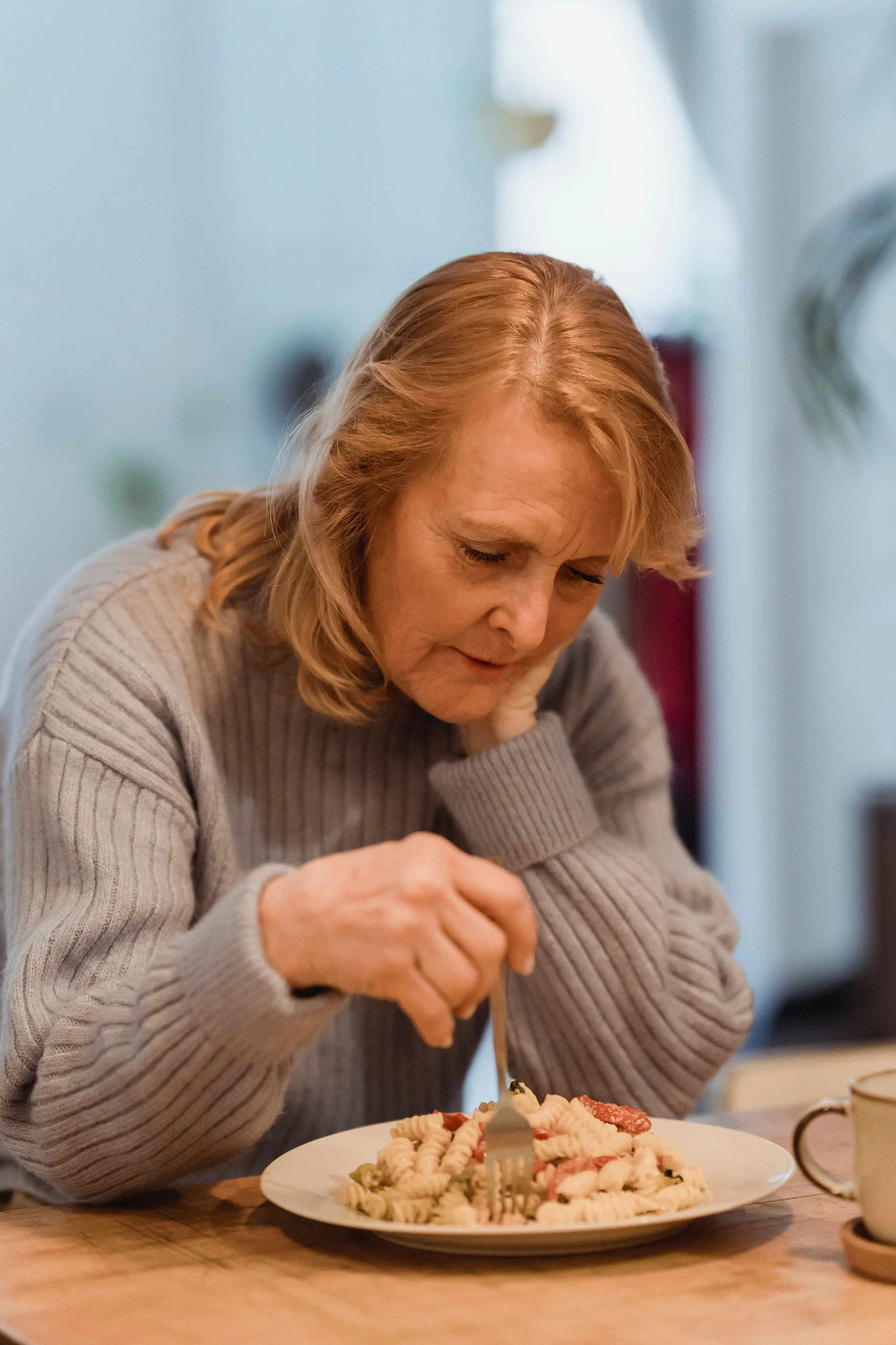 Une femme aux cheveux roux mange un repas à table, un plat de pâtes avec des morceaux de viande et de la sauce est devant elle.