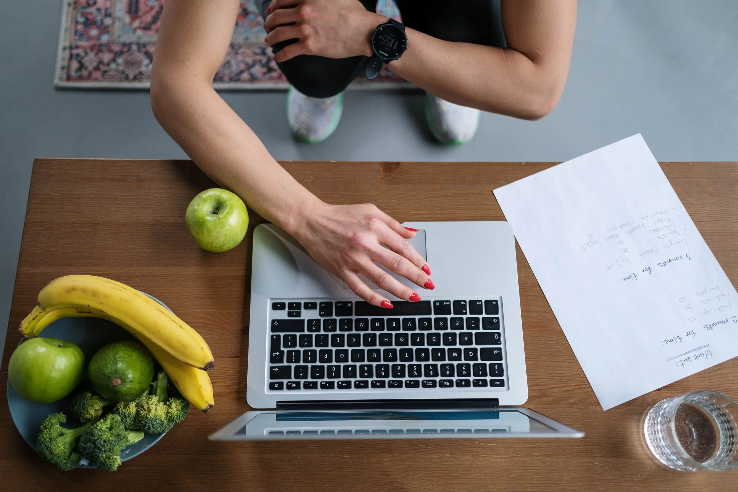Une diététicienne-nutritionniste faisant des consultations en visioconférence avec un ordinateur portable à une table avec des fruits, notamment des pommes, des bananes, des brocolis et des avocats, ainsi que des feuilles de papier avec des notes.