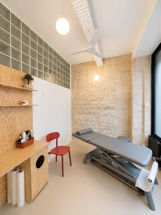 Une salle de consultation médicale avec une table d'examen, une chaise rouge, des étagères en bois, un ventilateur de plafond, et un mur en briques.