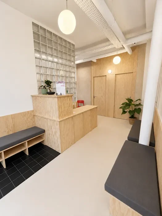 Salle d'attente avec un comptoir en bois, des bancs en bois avec coussins gris, une plante verte, une cloison en briques de verre, et un éclairage doux
