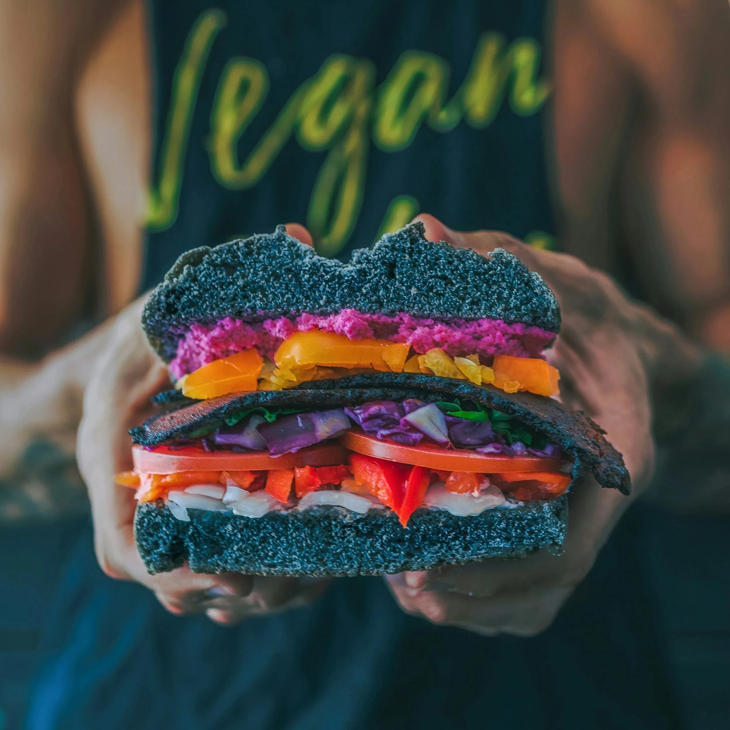 Un sandwich coloré avec plusieurs couches de légumes et de pain multicolore, tenu par une personne portant un t-shirt noir avec le mot 'Vegan' écrit en vert fluo.