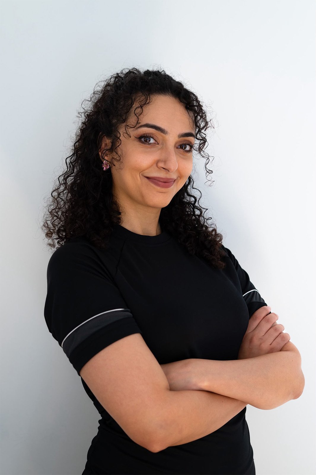 Jeune femme aux cheveux bouclés, portant un t-shirt noir, se tenant contre un mur blanc, souriant avec les bras croisés.