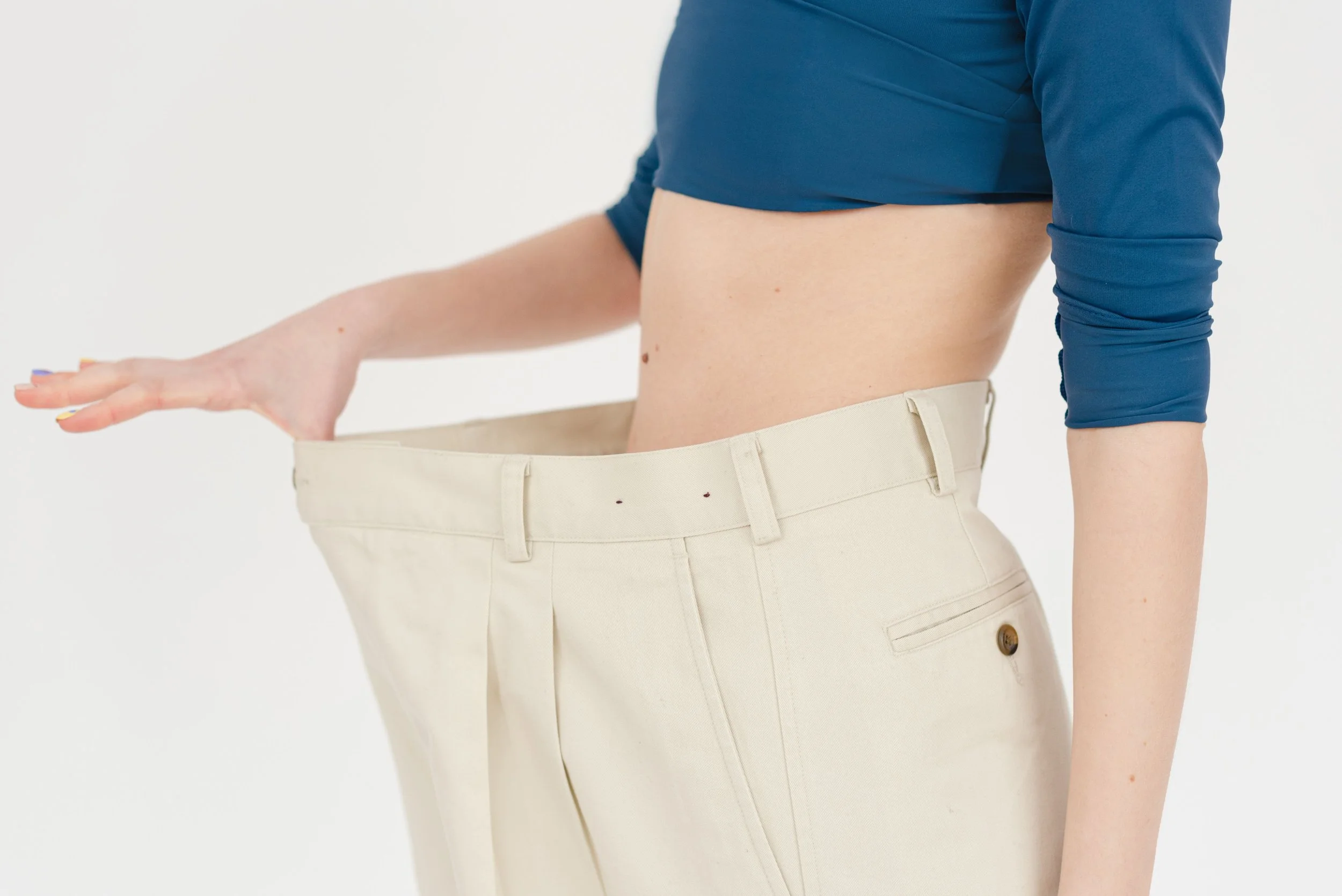 Une personne portant un pantalon beige et un haut bleu, tenant son pantalon pour montrer qu'il est trop grand, symbolisant une perte pondérale réussie, grâce à une prise en charge nutritionnelle personnalisée