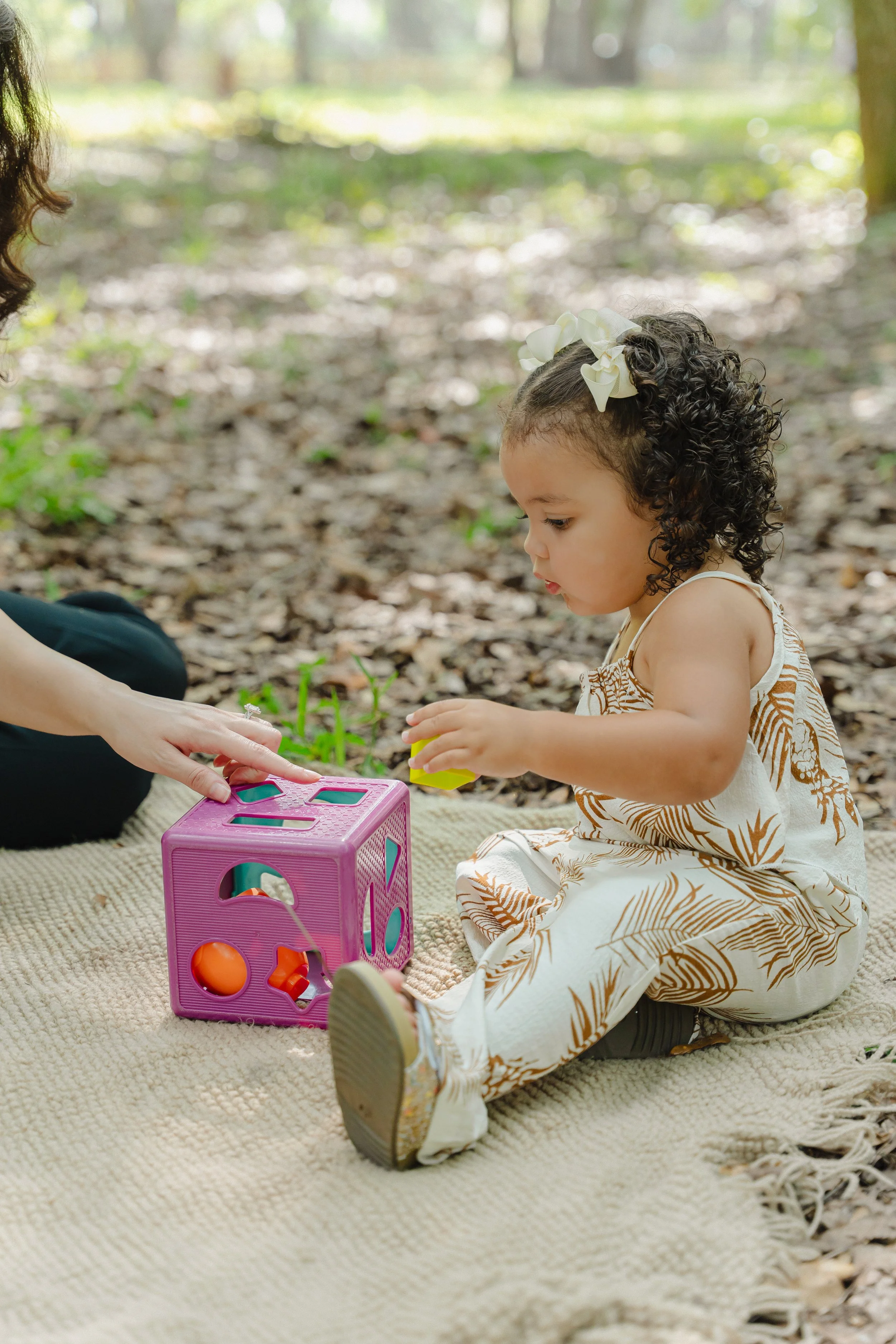 A young girl with curly hair and a white flower headband sitting on a beige blanket outdoors, playing with colorful toys under a tree surrounded by leaves and sunlight.
