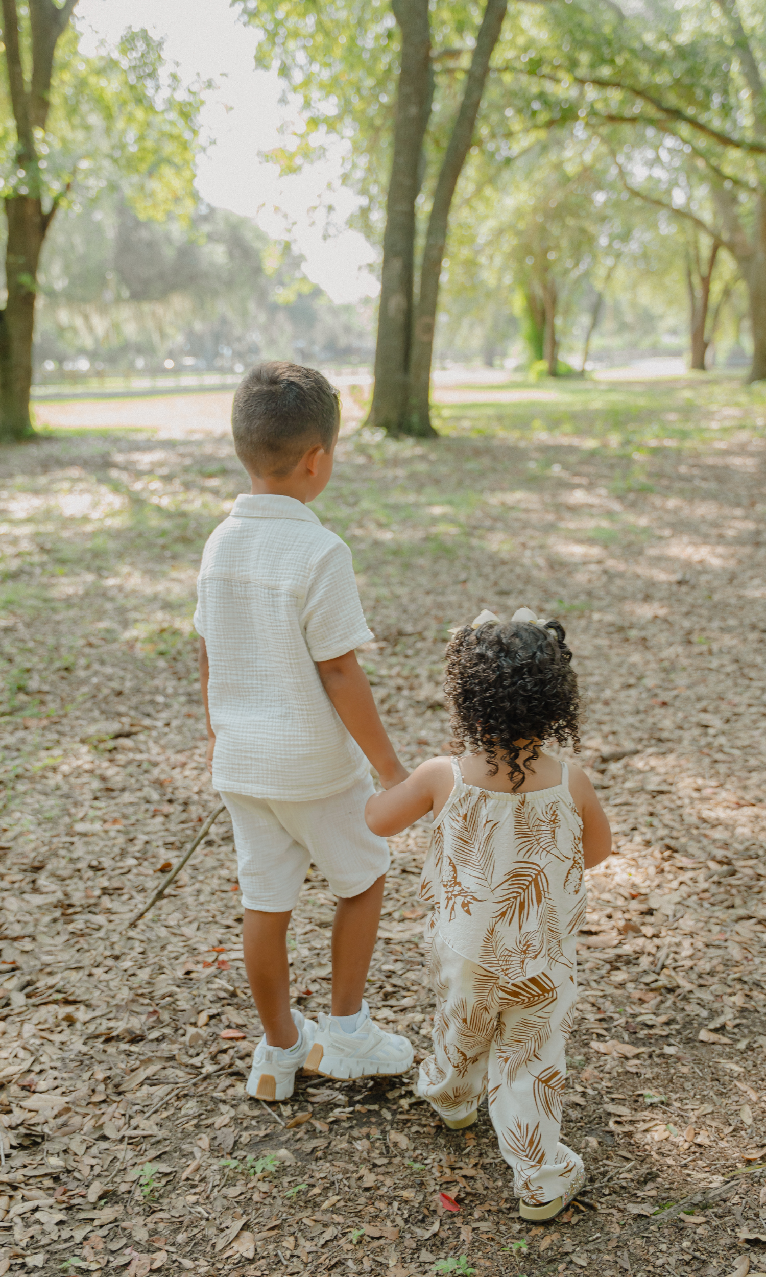 Two children, a boy and a girl, walking hand-in-hand on a wooded path in a park, surrounded by trees with sunlight filtering through the leaves.