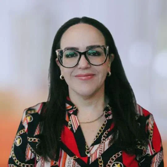 A woman with long dark hair wearing glasses, gold earrings, and a colorful patterned blouse with red, black, and gold accents, smiling at the camera against a neutral background.