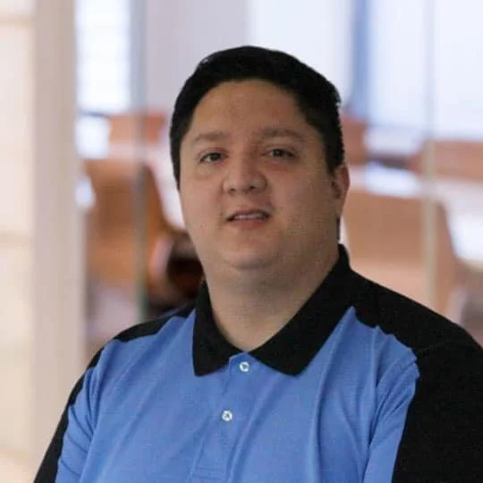 A man with short dark hair wearing a blue collared shirt with black accents, standing indoors with blurred background.